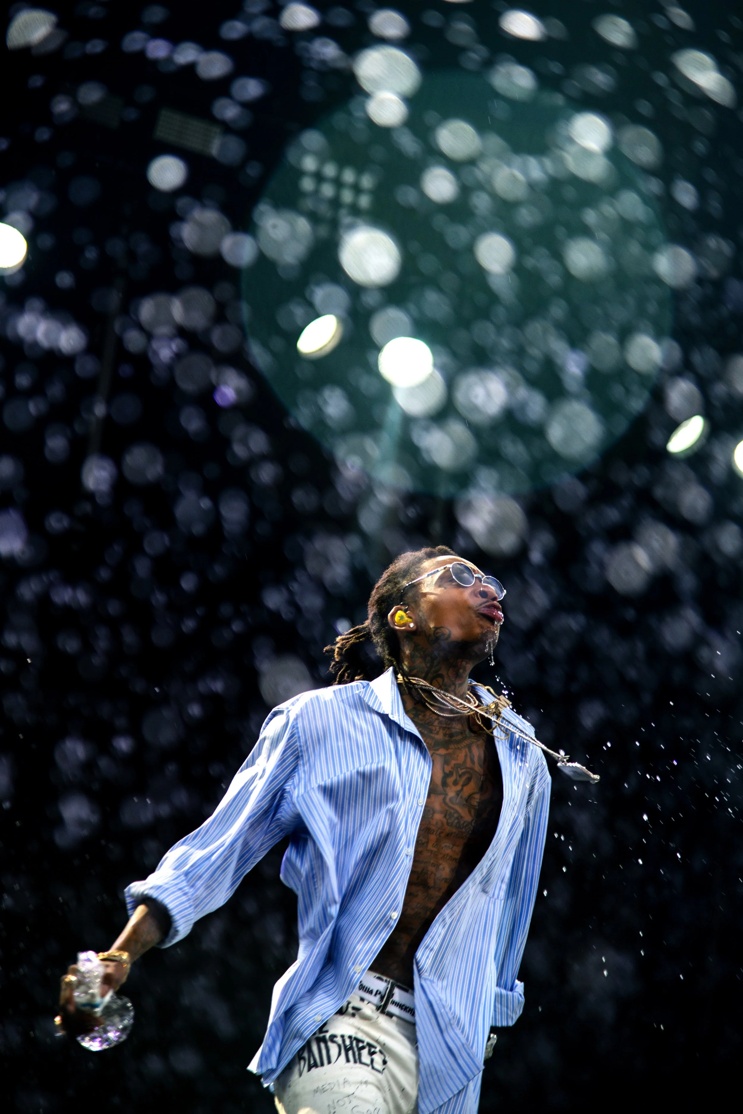 A male performer with tattoos on his chest, wearing sunglasses, a partially unbuttoned striped shirt, and white pants with black text, on stage with bright, dark background and bokeh lights.