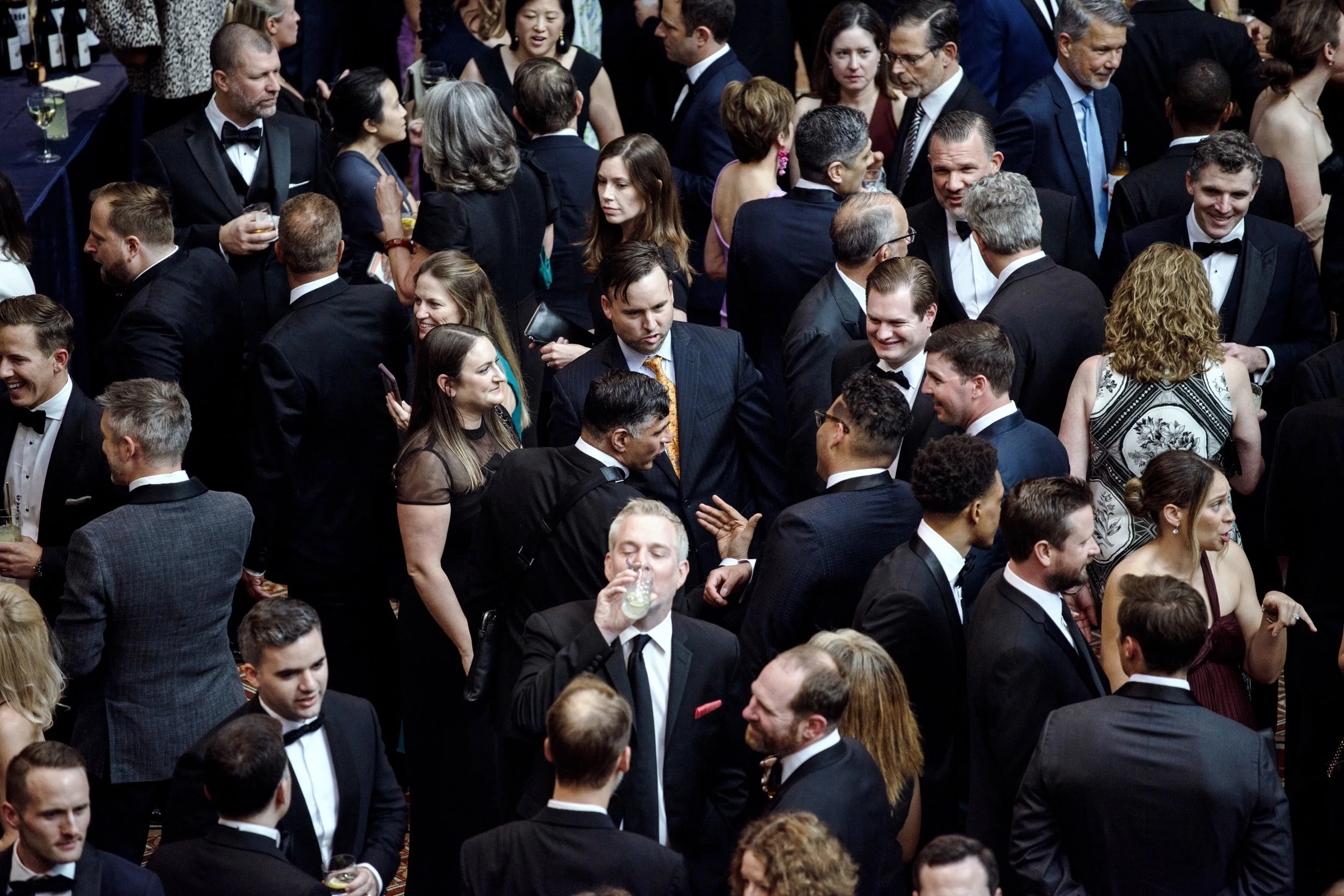 A large crowd of people dressed in formal attire at a social event or party, some holding drinks, mingling and talking.