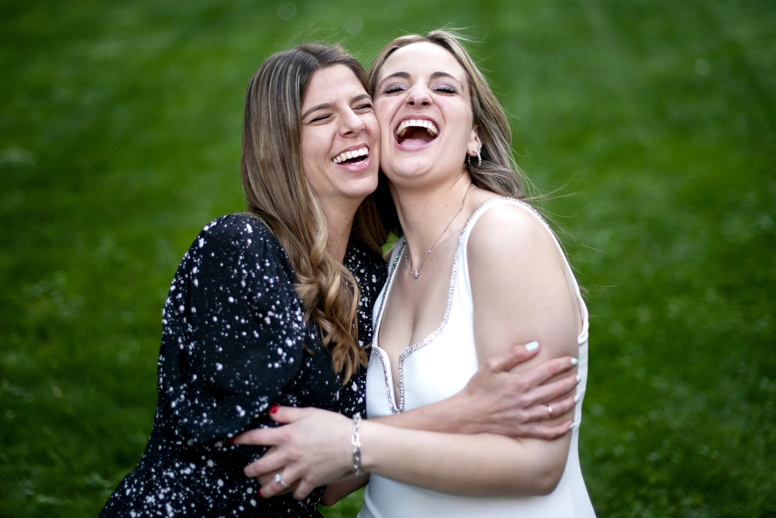 Two women hugging and laughing outdoors on a grassy field.