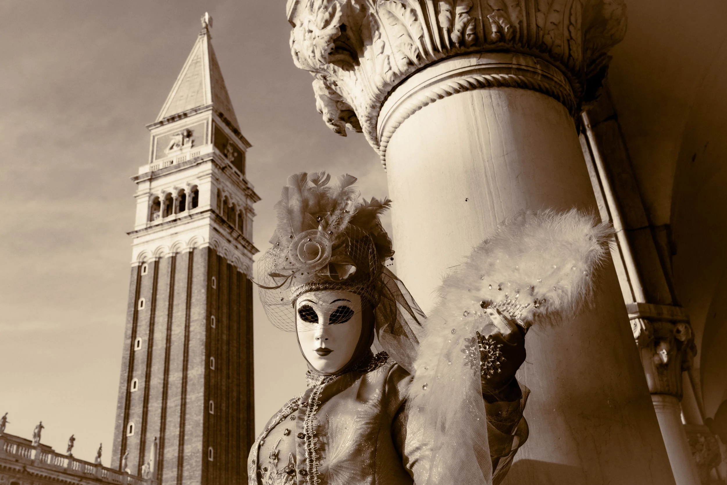 Venetian carnival mask with elaborate feathers, jewelry, and costume, set against a historic tower with a clock and a cloudy sky.