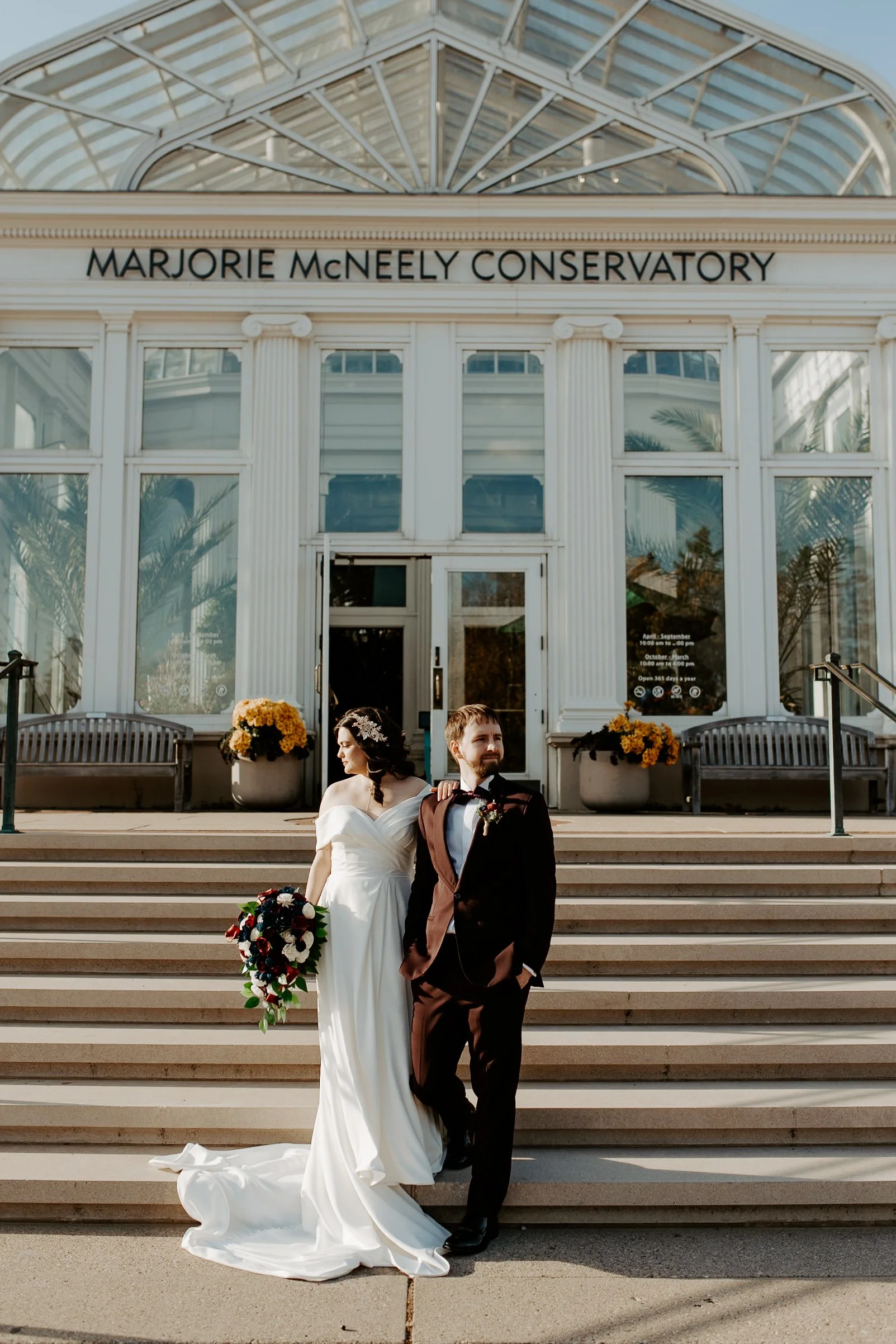 bride and groom standing in front of Como Zoo Conservatory in the fall with mums and a train and a bridal hair piece and the groom is wearing burgundy