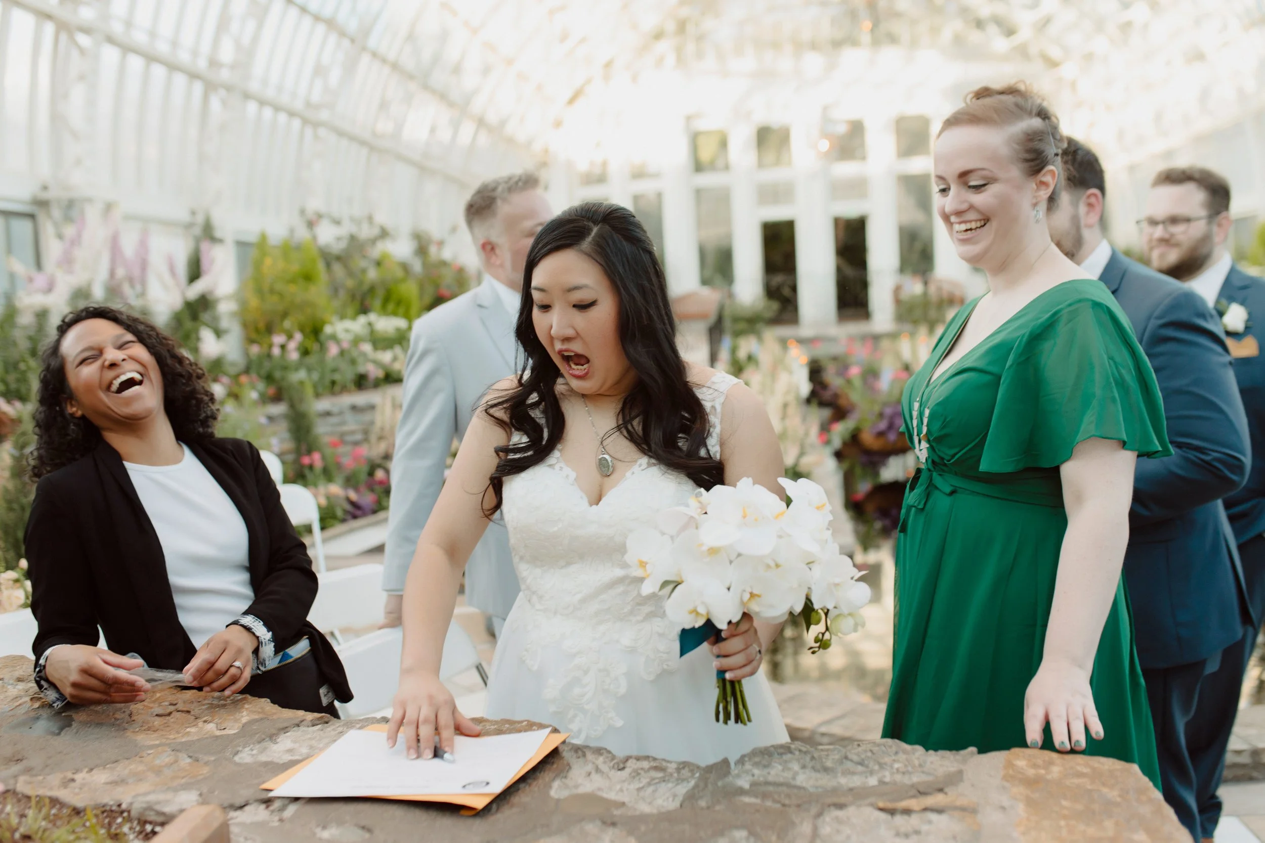 A bride holding a bouquet of white flowers and a man signing a document during a wedding ceremony in a greenhouse with sunlight, surrounded by smiling guests. Planner is laughing at the bride's joke
