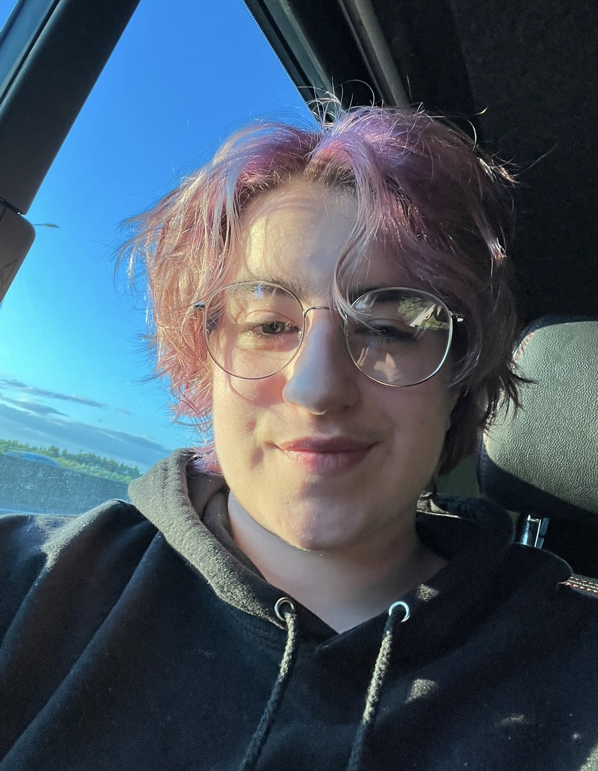 Kilbo has short pink hair and round wire-framed glasses. He is wearing a black hoodie and has a neutral expression