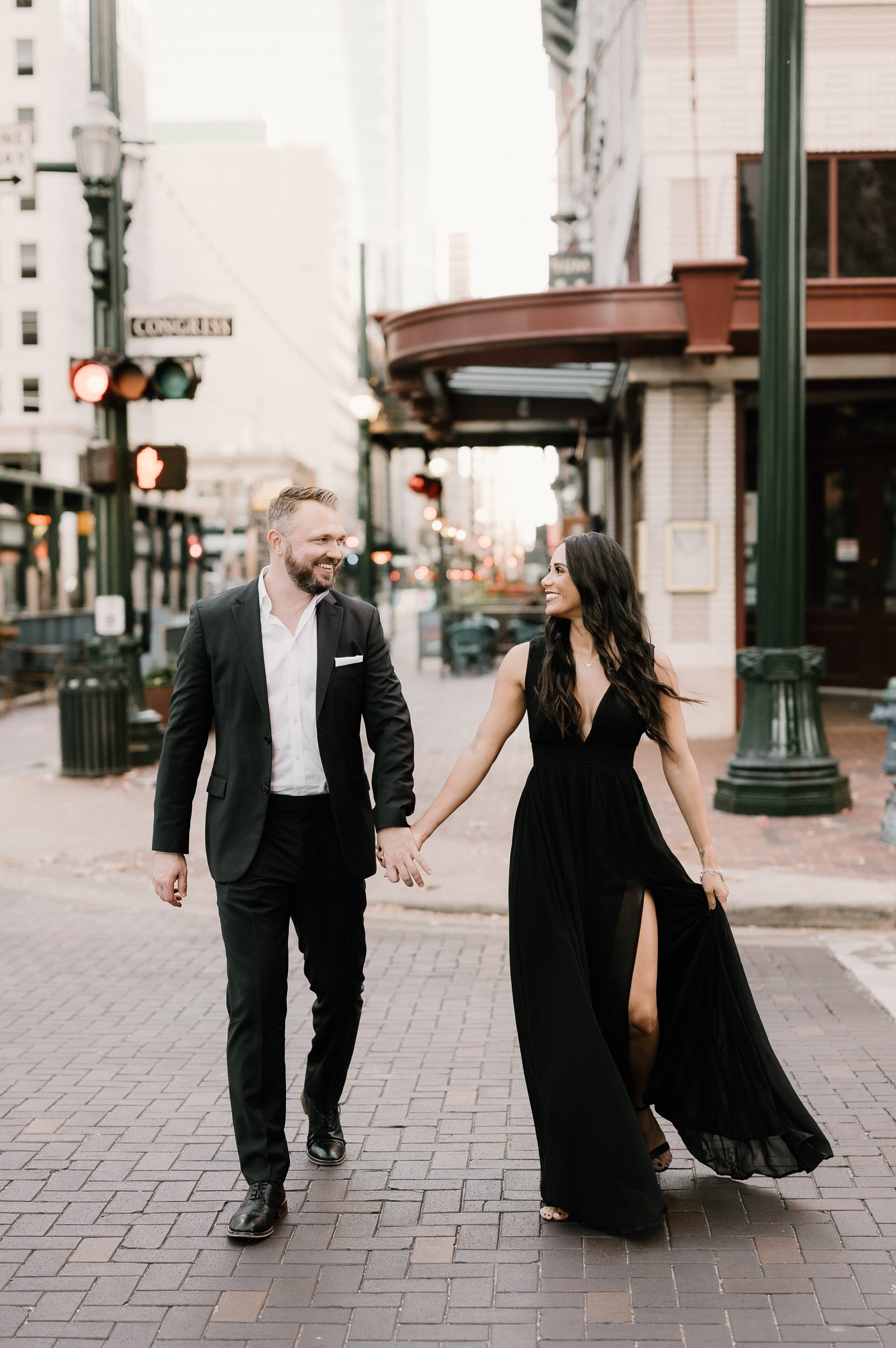 A couple walking hand-in-hand in an urban street, dressed formally, with city buildings in the background.