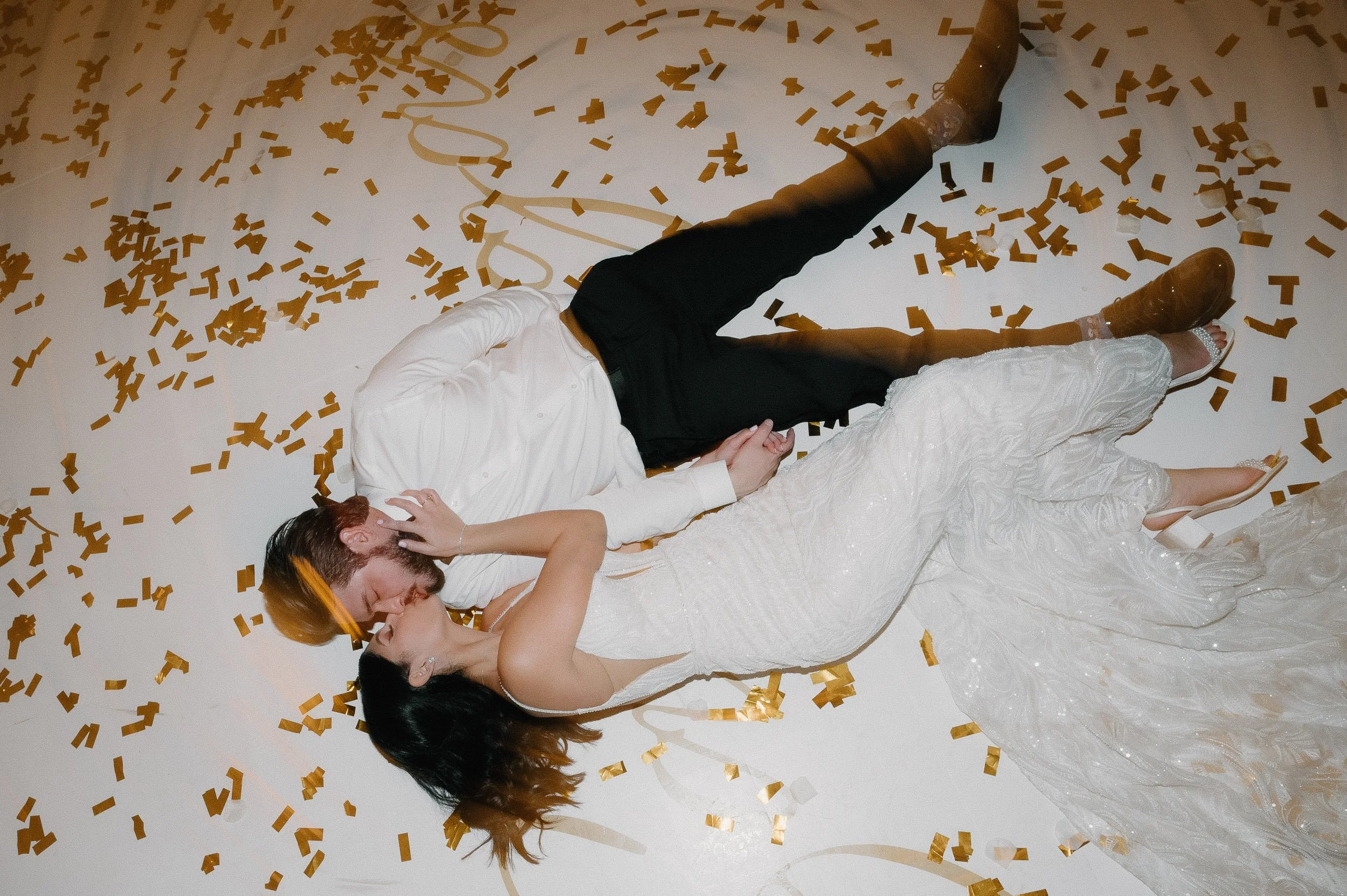 A couple lies on the floor, surrounded by gold confetti, with the man in a white shirt and black pants and the woman in a white wedding gown, sharing a kiss.