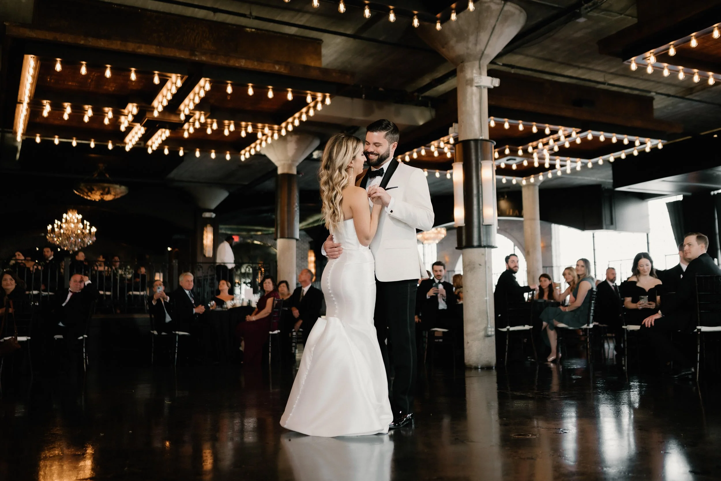A bride and groom sharing a dance in a reception hall with dim lighting, string lights hanging from the ceiling, and guests seated at tables watching.