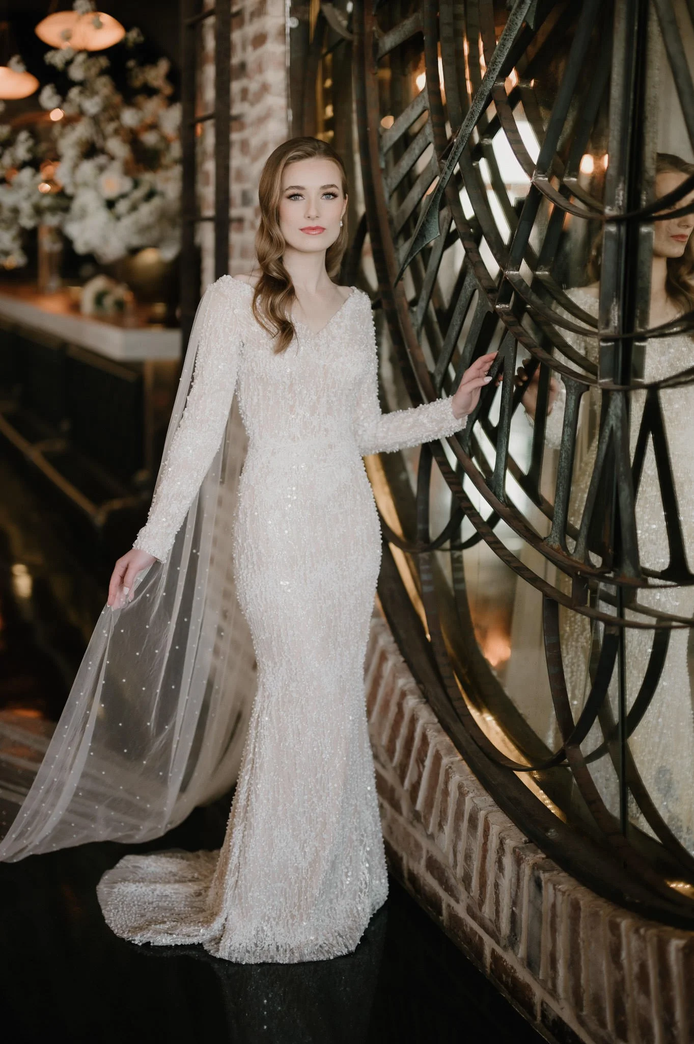 A woman in a long, elegant, white sequin wedding dress stands next to a large decorative mirror, with a blurred background of a decorated interior space.