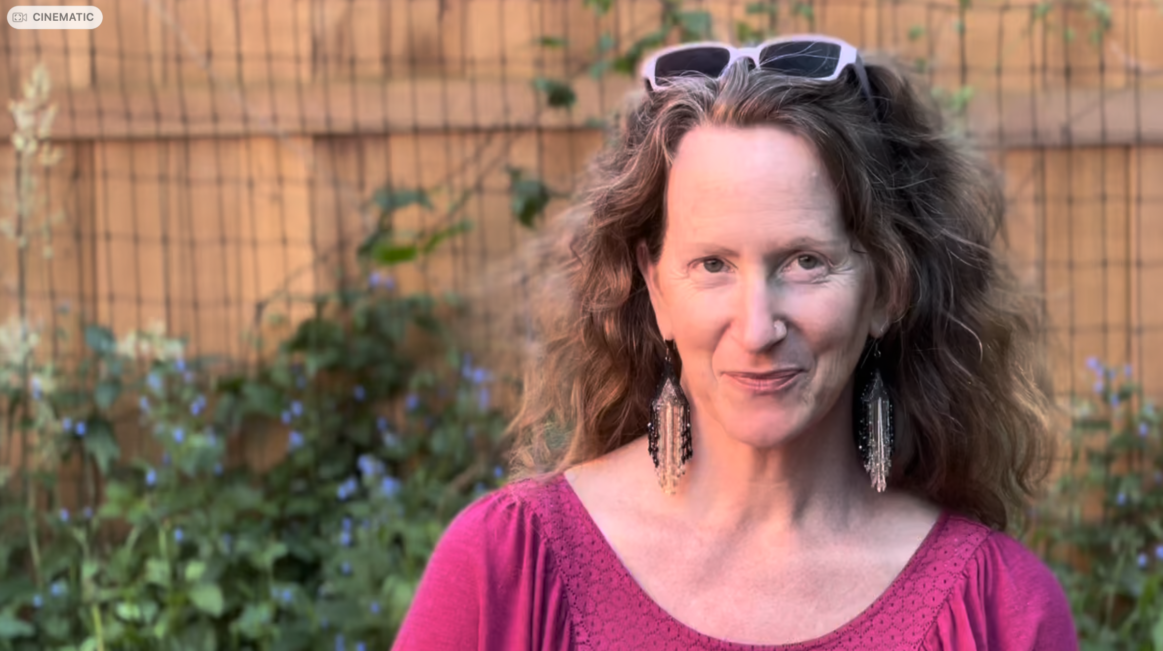 A woman with curly brown hair, wearing a pink lace top, large earrings, and sunglasses on her head, standing outdoors in front of a wooden fence with green plants.