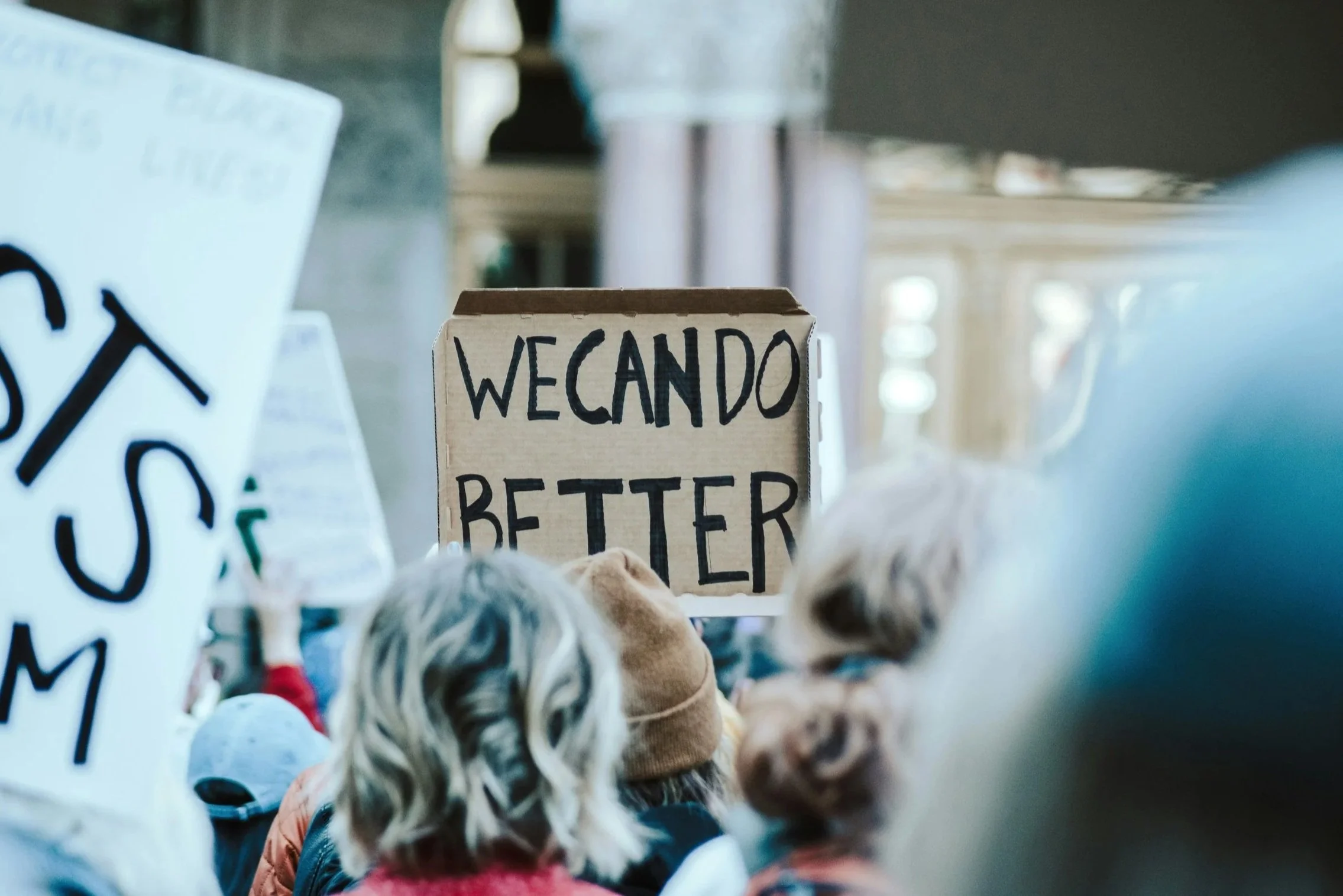 Person holding a handmade protest sign reading “WE CAN DO BETTER” above a crowd of people