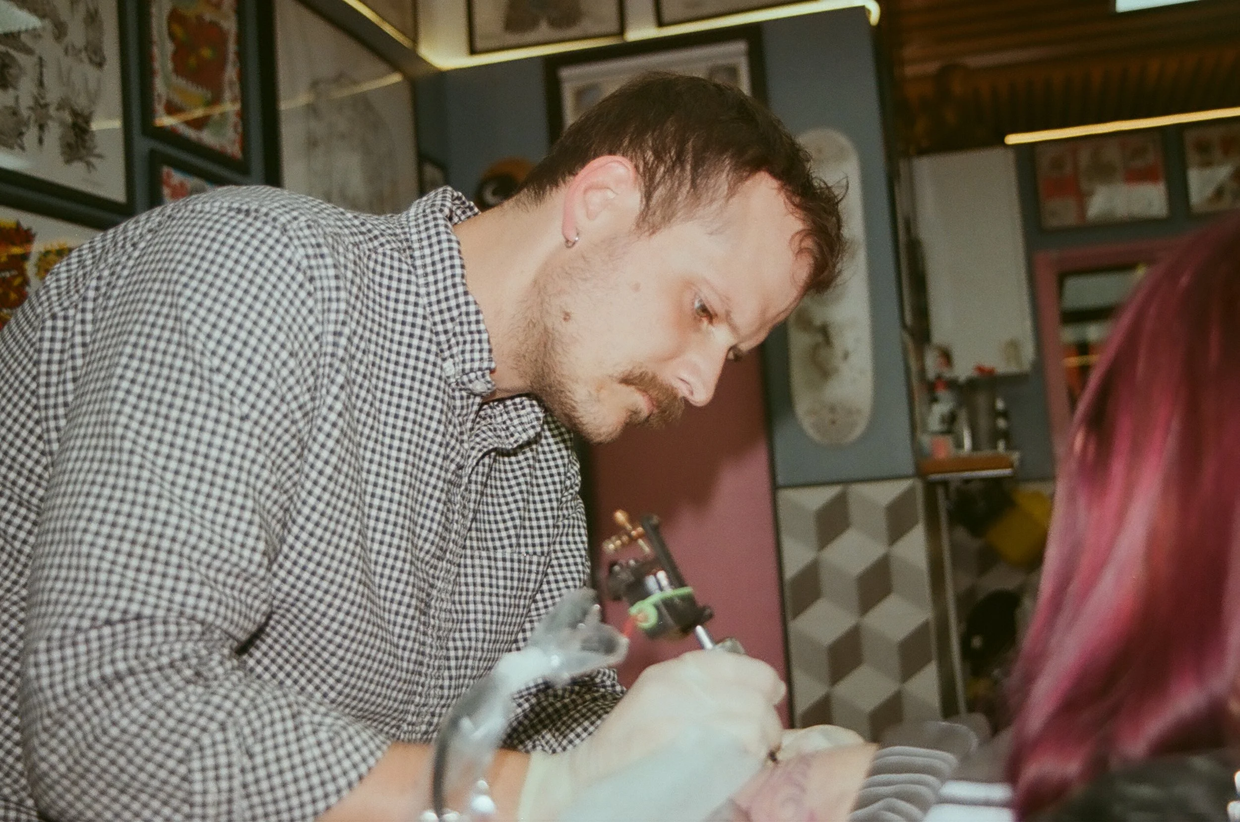 A man with a checkered shirt tattooing a person's arm in a tattoo studio.