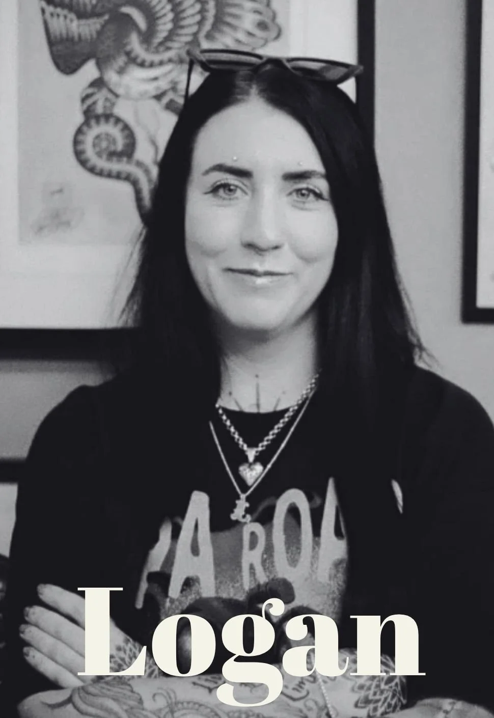 Logan the piercer at Kilburn Original with dark hair and light skin smiling, wearing glasses on her head and layered necklaces, standing in front of framed artwork on a wall. The word 'Logan' is overlaid at the bottom.