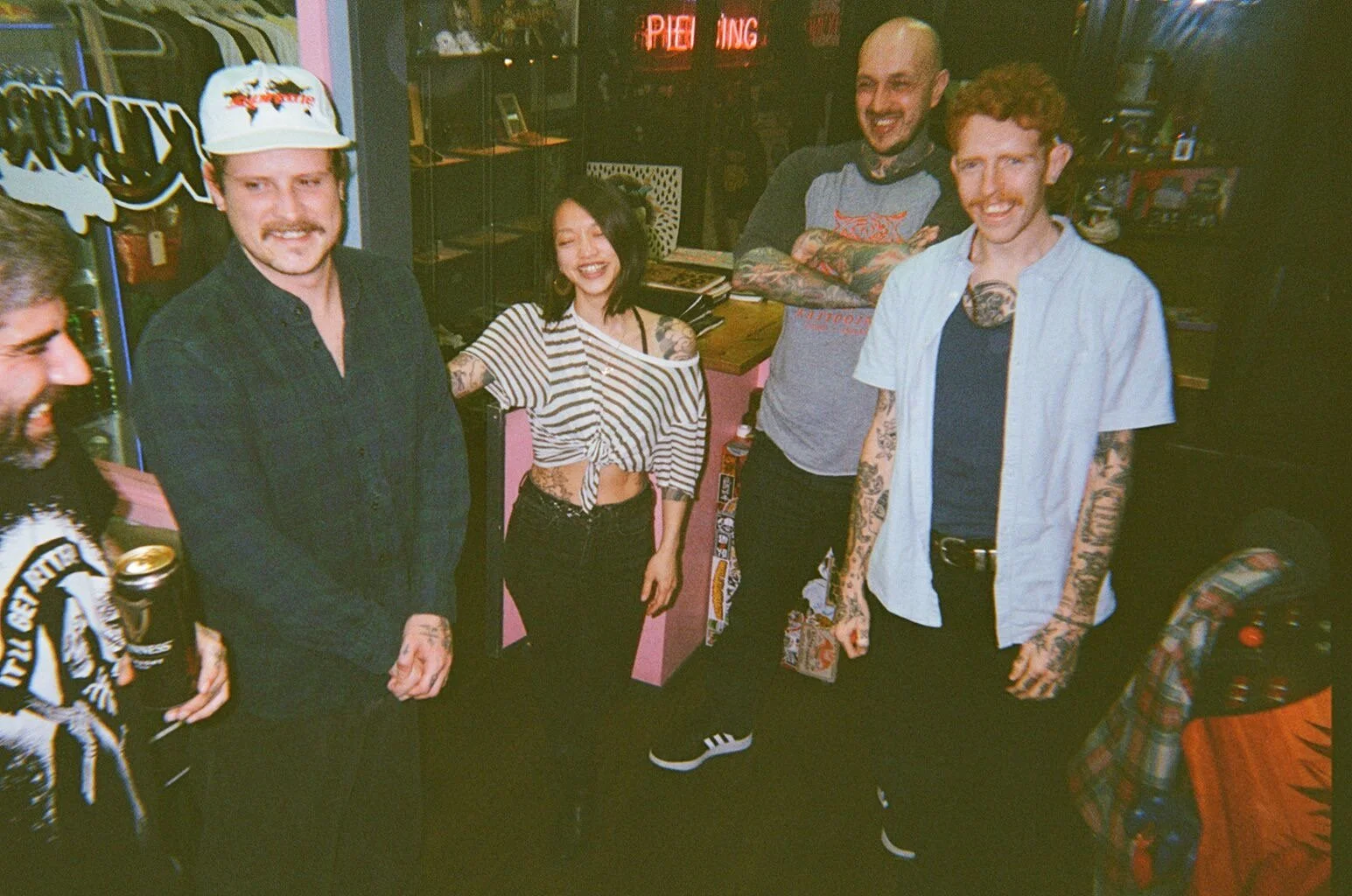 Group of five young adults smiling and posing in a casual indoor setting, with neon signs in the background and some people holding drinks.