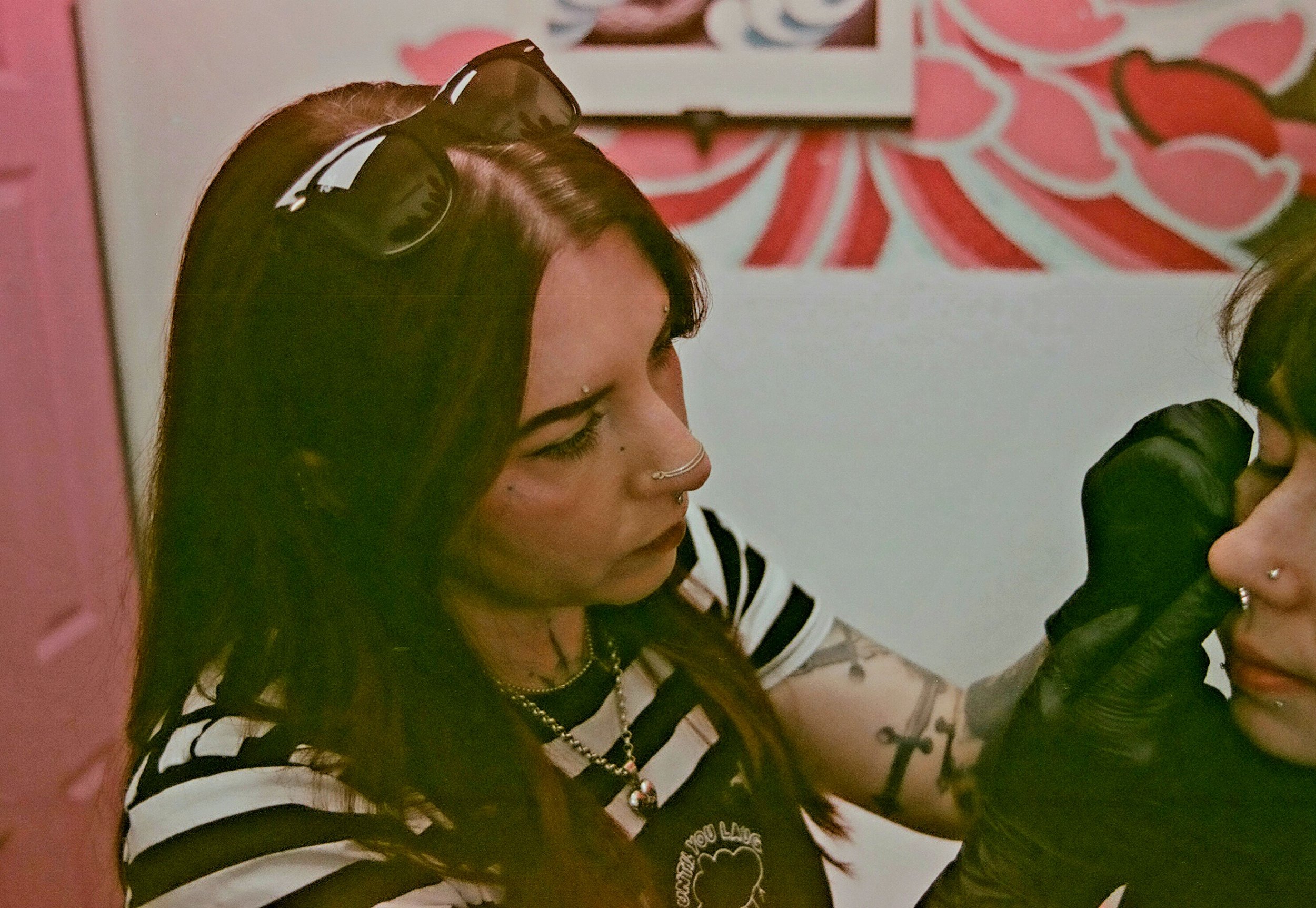 Piercer Logan with long, dark hair, braided at the end, wearing sunglasses on her head, a nose ring, and a black and white striped shirt with a necklace, performing an ear piercing with a needle