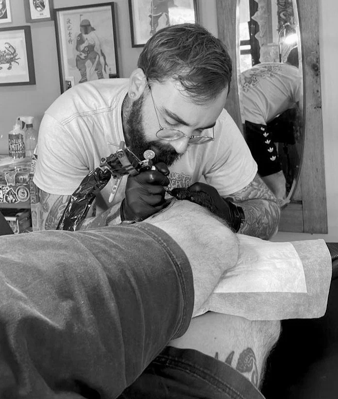 A tattoo artist working on a client's leg in a tattoo studio, with framed art and a mirror visible in the background.