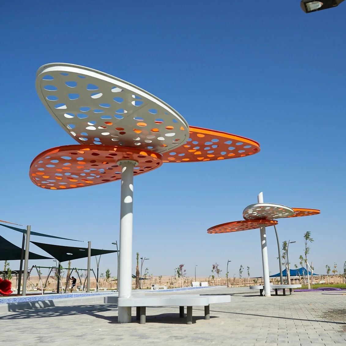 פרגולות אזימוט במתכת כתומה ולבנה מחוררת על עמודים לבנים, מרחב ציבורי מרוצף בשכונת השחר דימונה