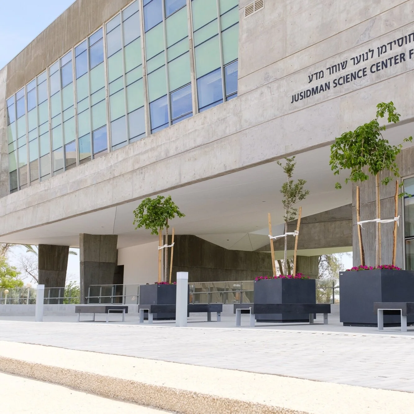 מבט חזיתי על כניסת מרכז חוסידמן למדע לנוער עם מיכלי שתילה ממתכת וספסלי בטון