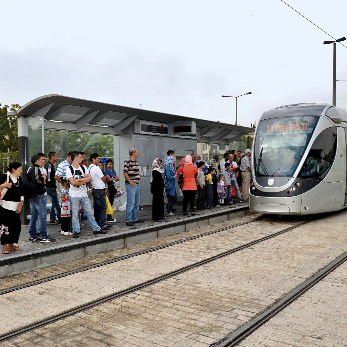קבוצת אנשים ממתינה בתחנת רכבת קלה ירושלים לצד פביליון המתנה ממתכת וזכוכית