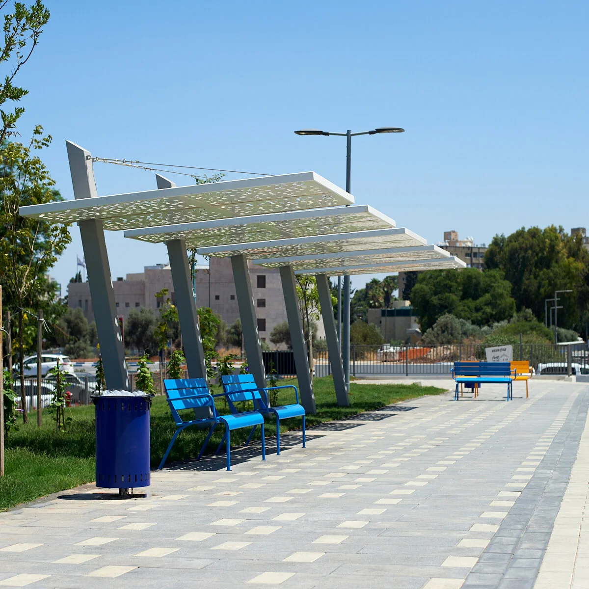 כיסאות מתכת צהובים וספסל כחול לאורך שביל מרכז מסחרי תחת פביליון הצללה