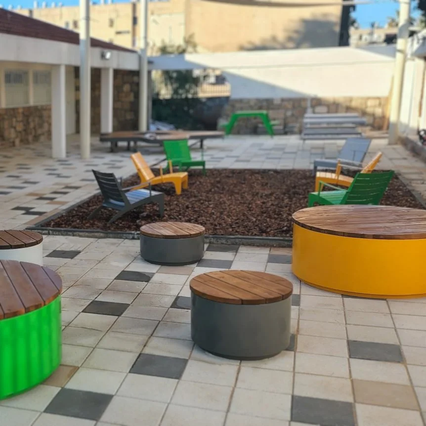 כורסאות חוץ בצבעי צהוב, ירוק ואפור על שבבי עץ, פביליון הצללה בחצר אולפנה