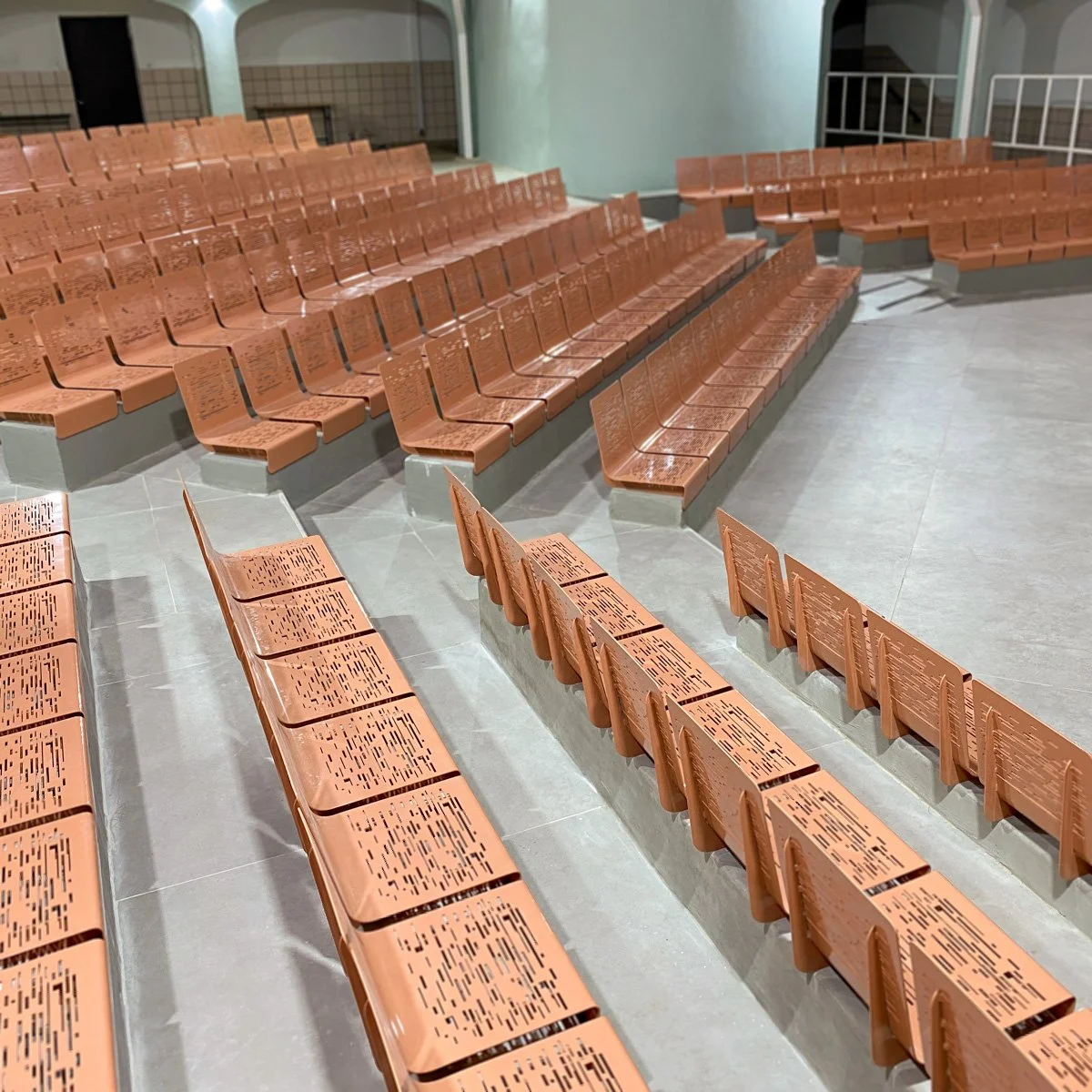 ספסלי מתכת בגוון טרקוטה עם גיזור לייזר גיאומטרי, שורה רציפה באודיטוריום מקורה