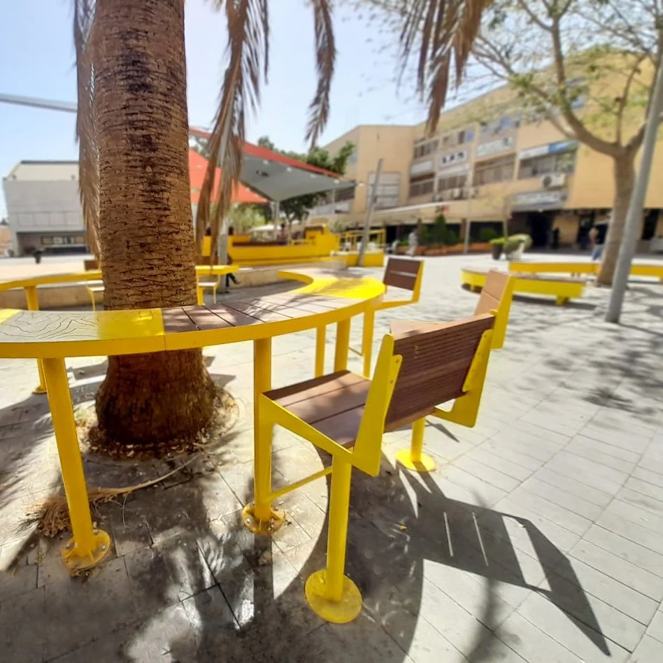 כיסאות מתכת צהובים עם מושב עץ כהה בכיכר ציבורית בערד, בניין רב קומות ברקע
