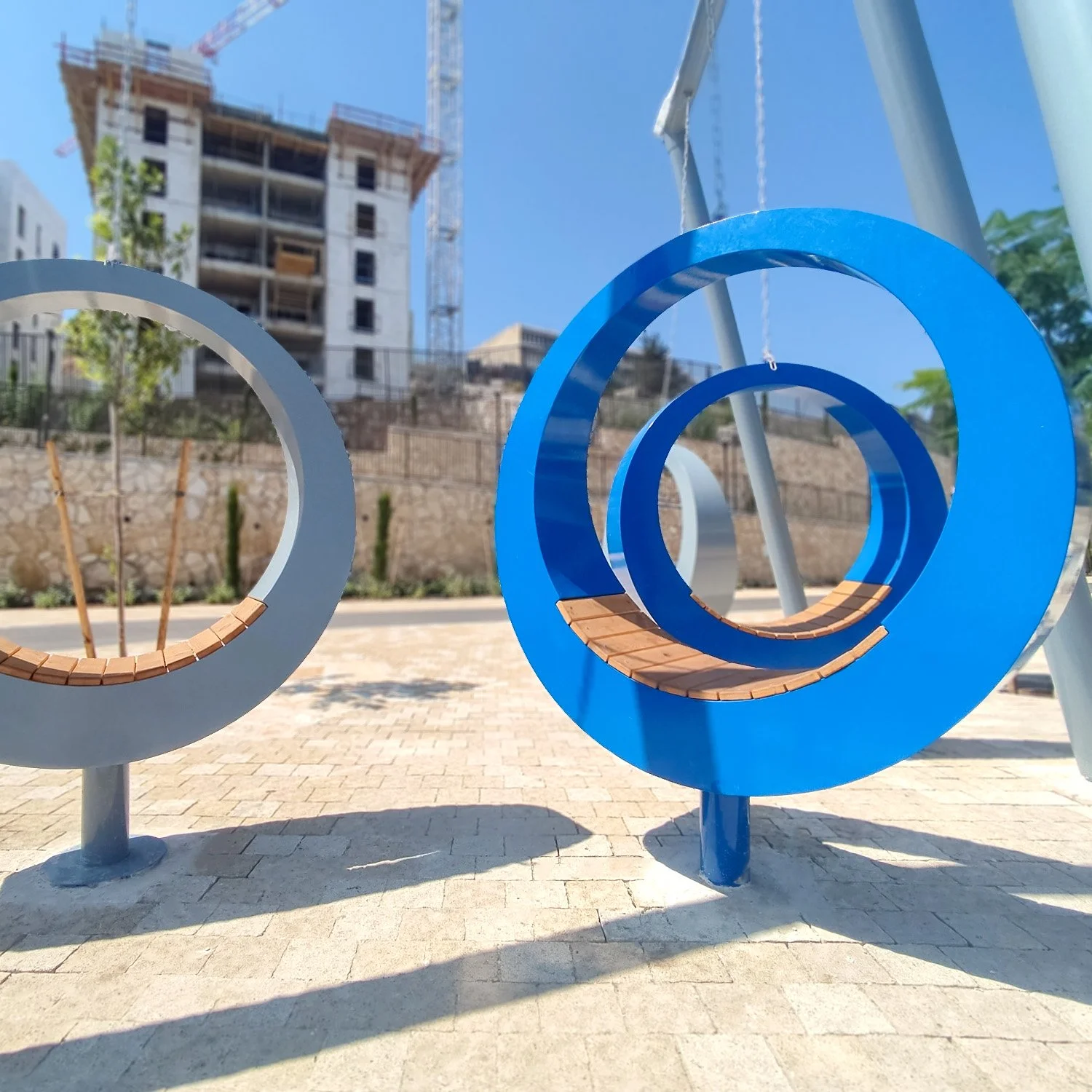 סדרת אלמנטי ישיבה פיסוליים – טבעות מתכת כחולות ואפורות עם מושבי עץ בפארק