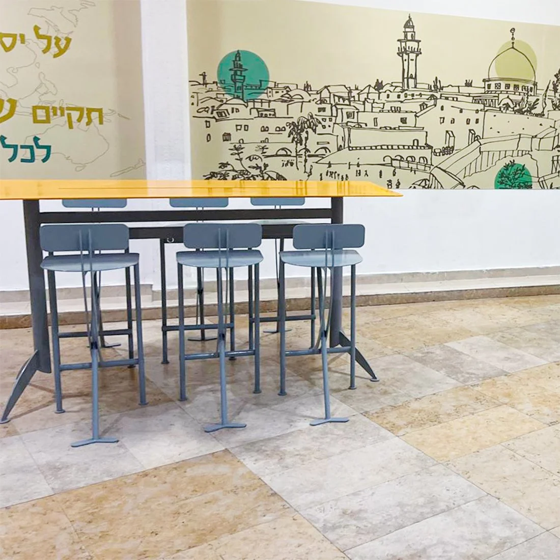שולחנות גבוהים עם משטח זכוכית צהובה וכיסאות בר מתכת אפורים, מסדרון עם ציורי קיר ירושלמיים