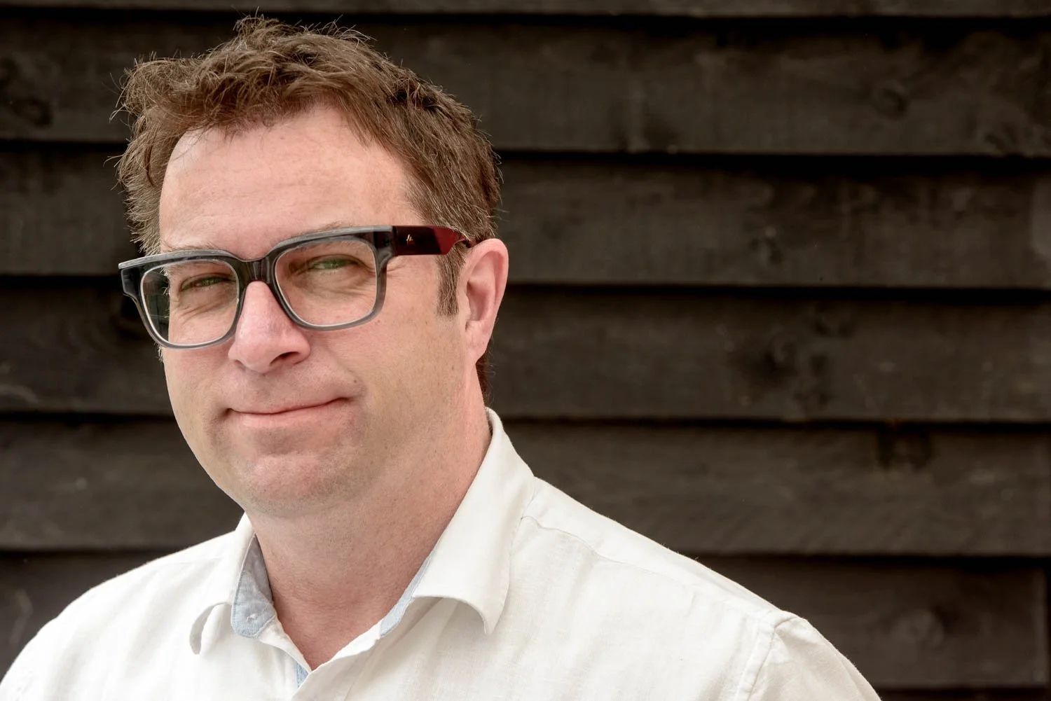 Richard Stonehouse, Architectural and built environment photographer, wearing glasses and a white collared shirt, standing outdoors in front of a dark wooden wall.