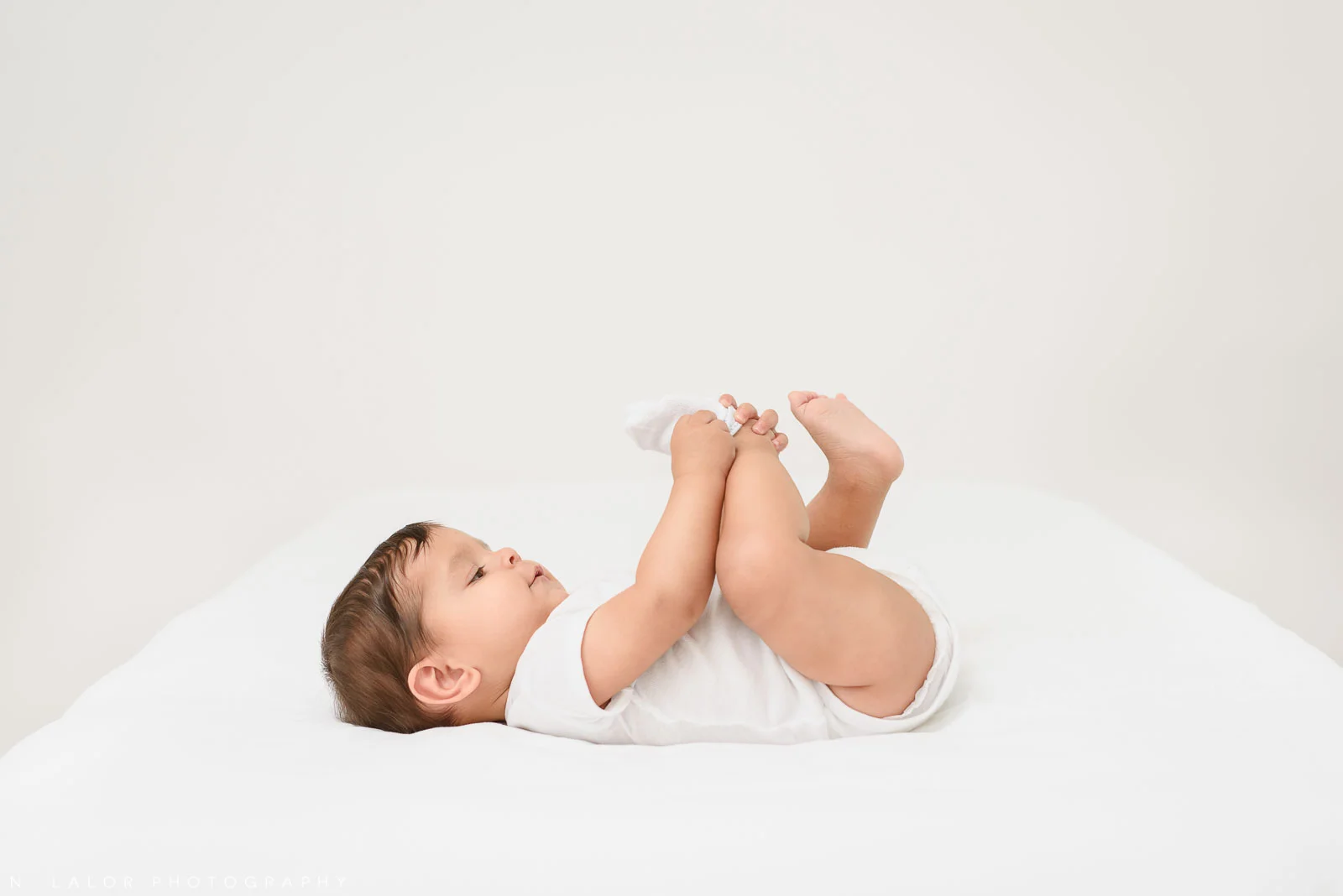Image of baby in white shirt playing with toes during photo shoot. Milestone Sessions by N. Lalor Photography. Studio in Greenwich, Connecticut.