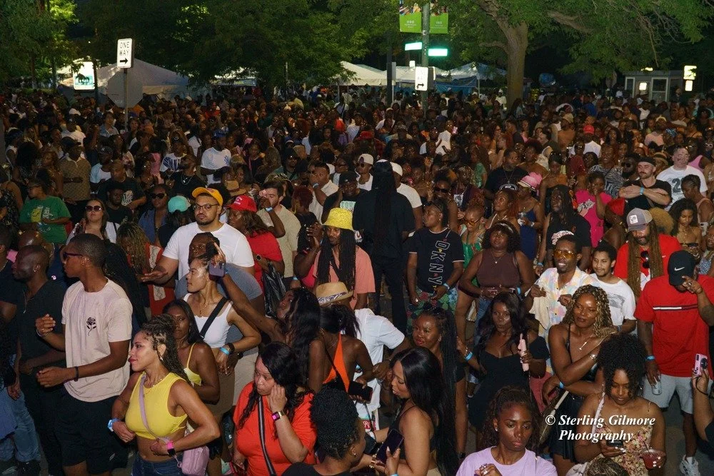 Large crowd of people gathered outdoors at night, some wearing hats and sunglasses, with trees and tents in the background.