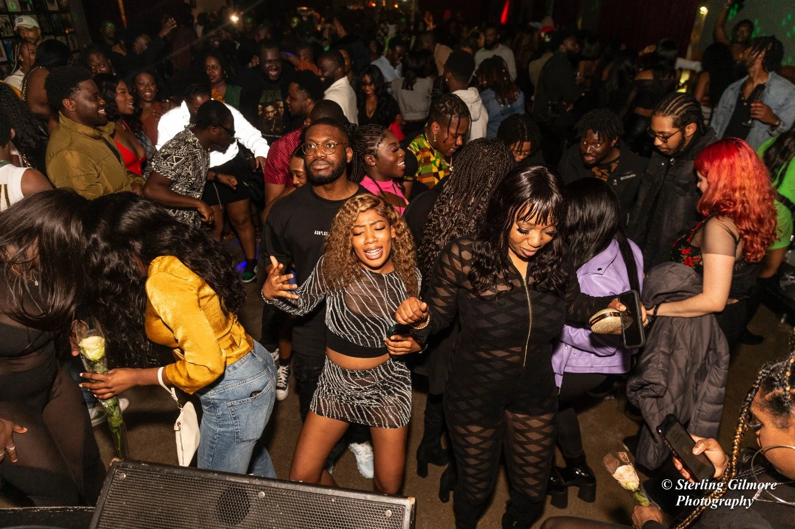 A crowded indoor party with people dancing and socializing, some holding flowers and taking photos.