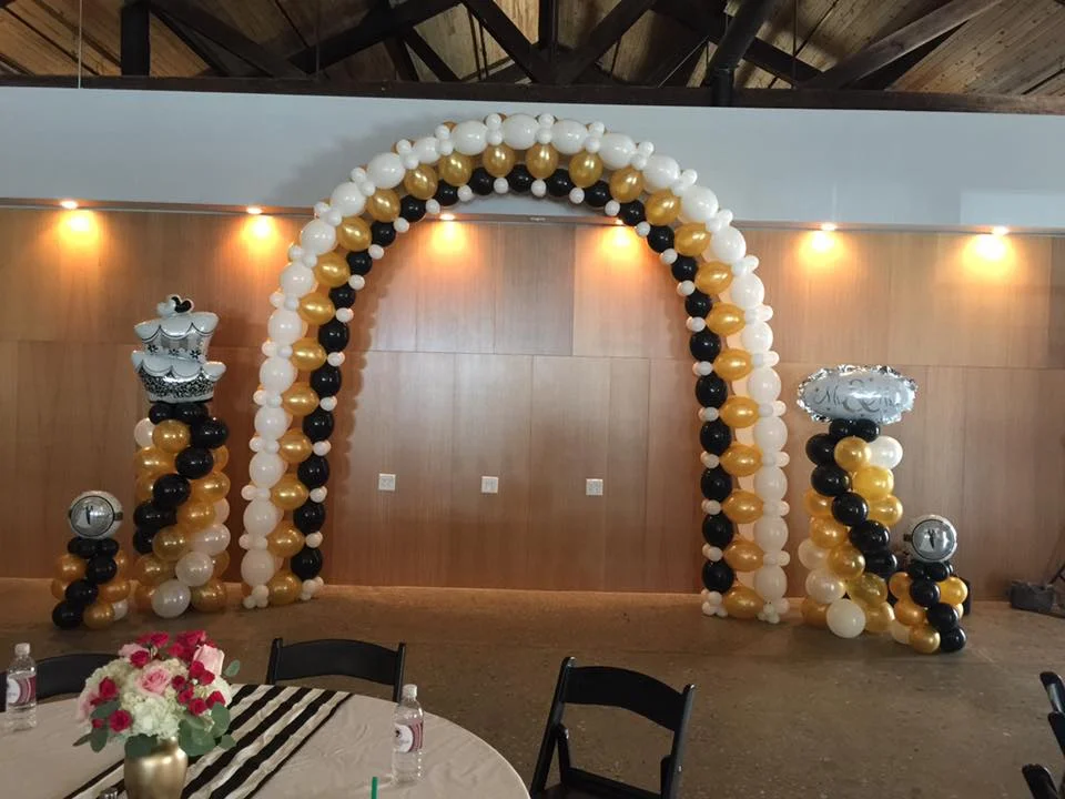 Balloon arch and columns decorated for a celebration, featuring a large arch made of white, gold, and black balloons, with two columns on either side decorated with similar balloons, topped with themed balloon arrangements.