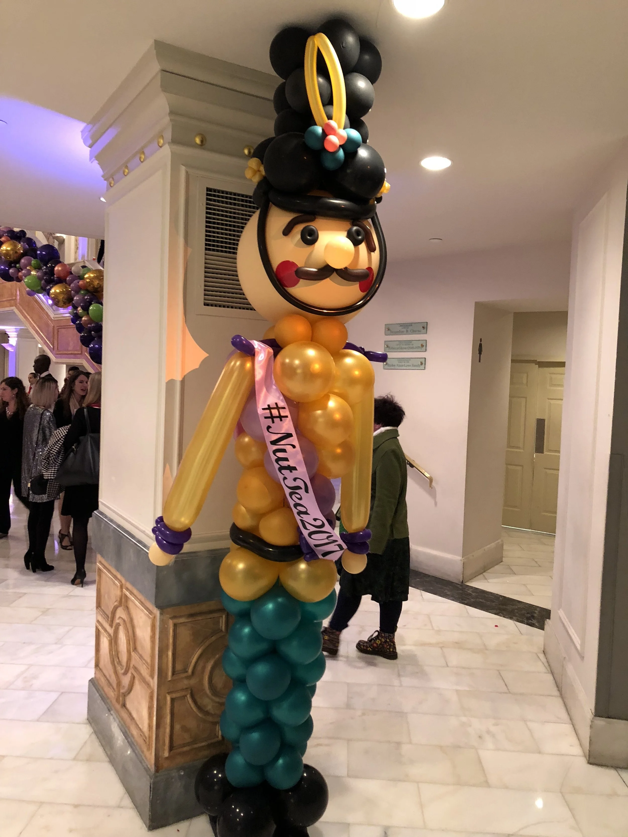 Balloon sculpture of a tall, bearded nutcracker soldier, with a black hat, yellow beard, and colorful clothing, wearing a sash with the hashtag #Yucat2022.