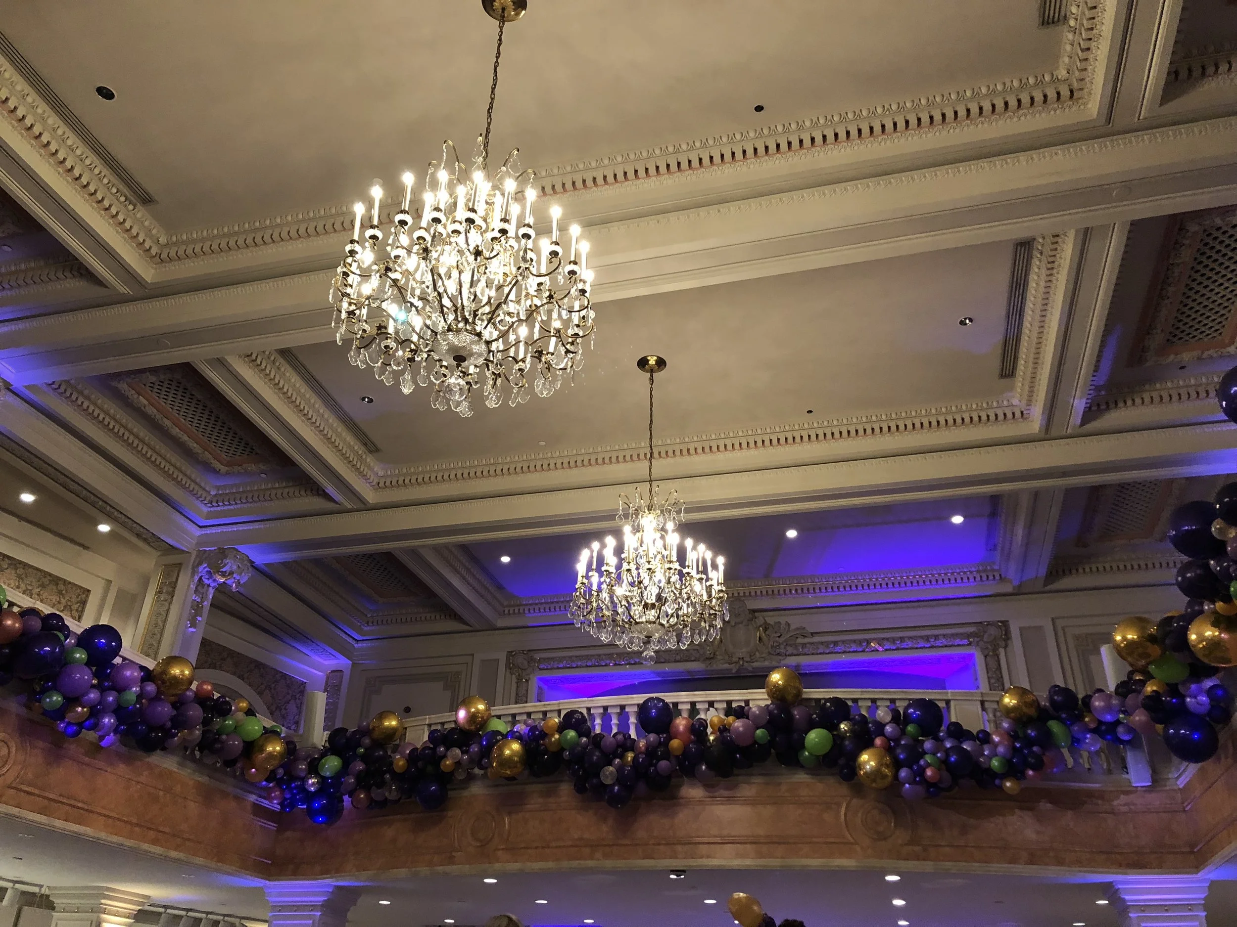 Elegant room interior with ornate ceiling, two large chandeliers, and a decorated balcony with black, purple, gold, and green balloons.
