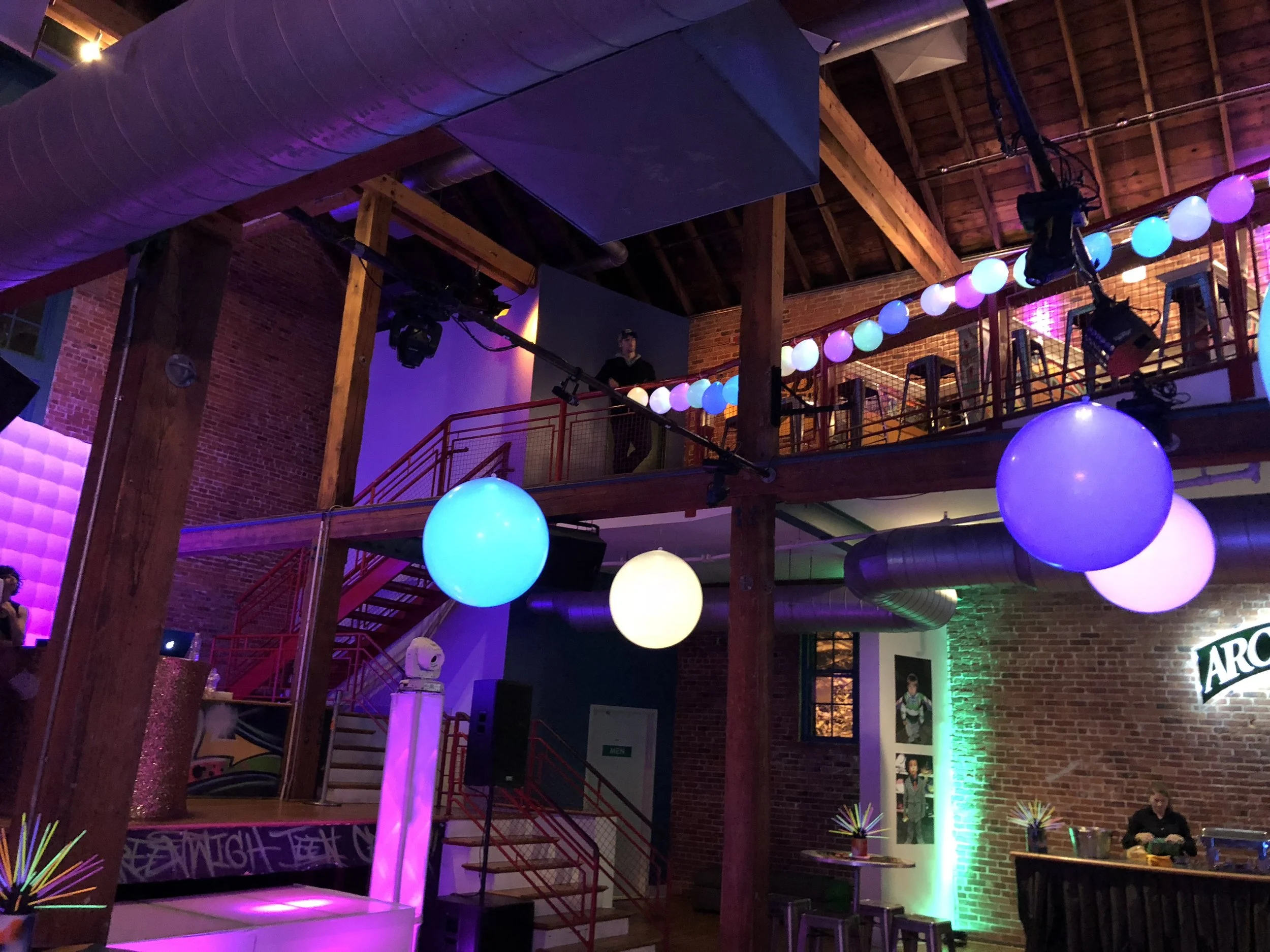 Interior of a lively bar or club with colorful LED, hanging balloons, a staircase, and a brick wall with artwork.
