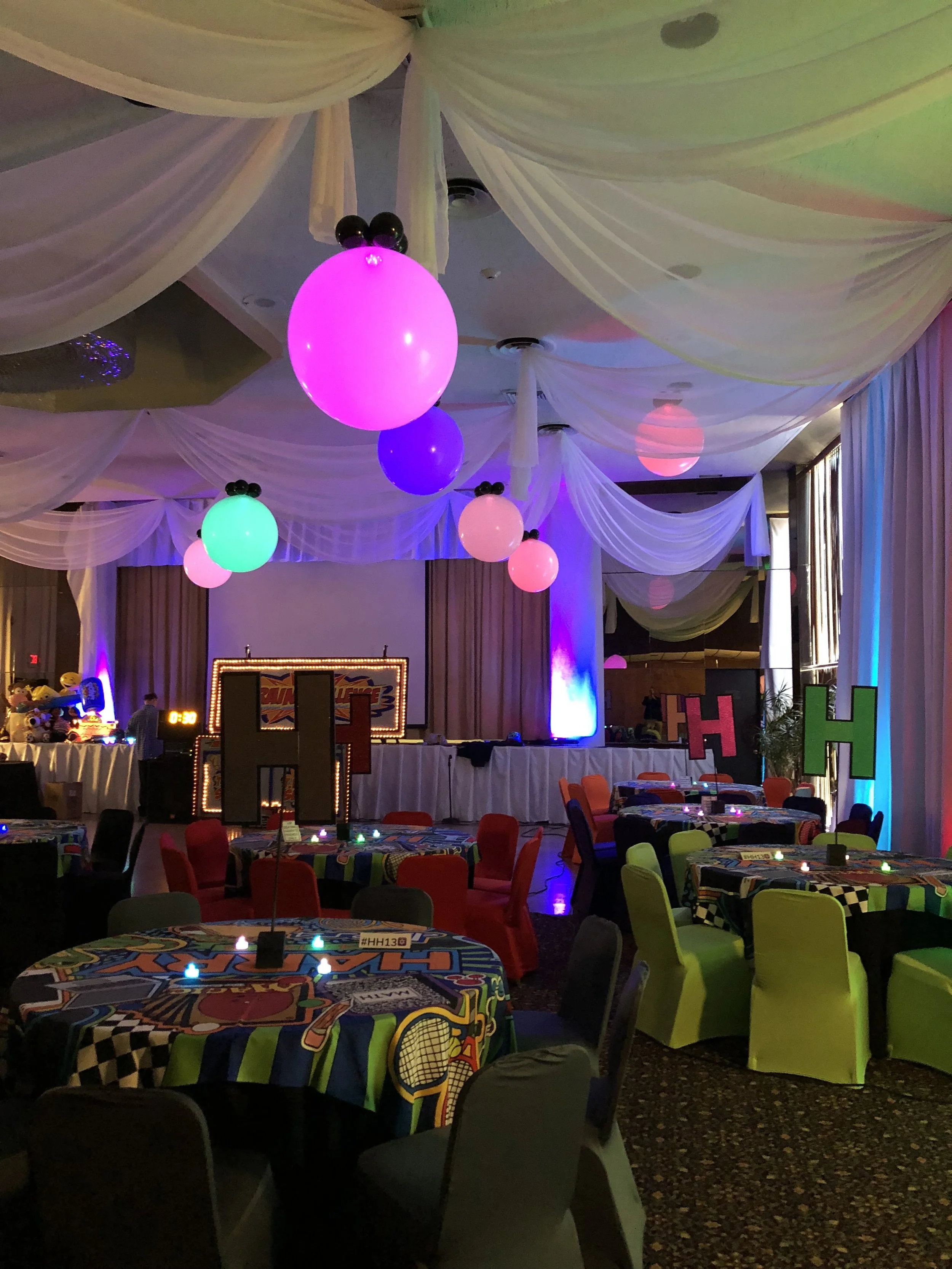 Colorful decorated party room with brightly colored tables, chairs, and hanging LED balloons lights in pink, purple, blue, and green, with a stage in the background.