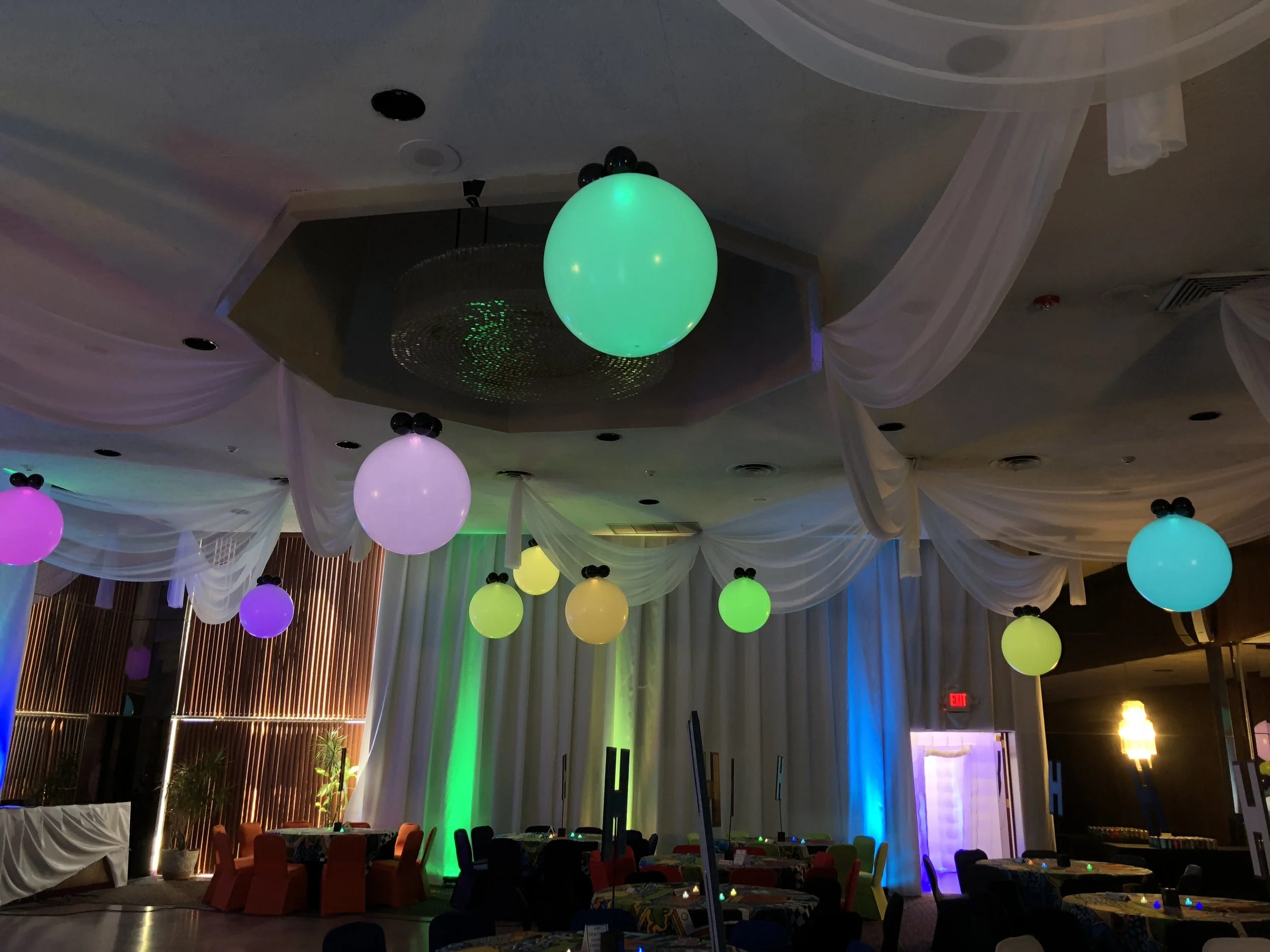 Interior of a decorated event space with colorful LED balloons hanging from the ceiling and draped fabric, illuminated by various lights including green and purple.