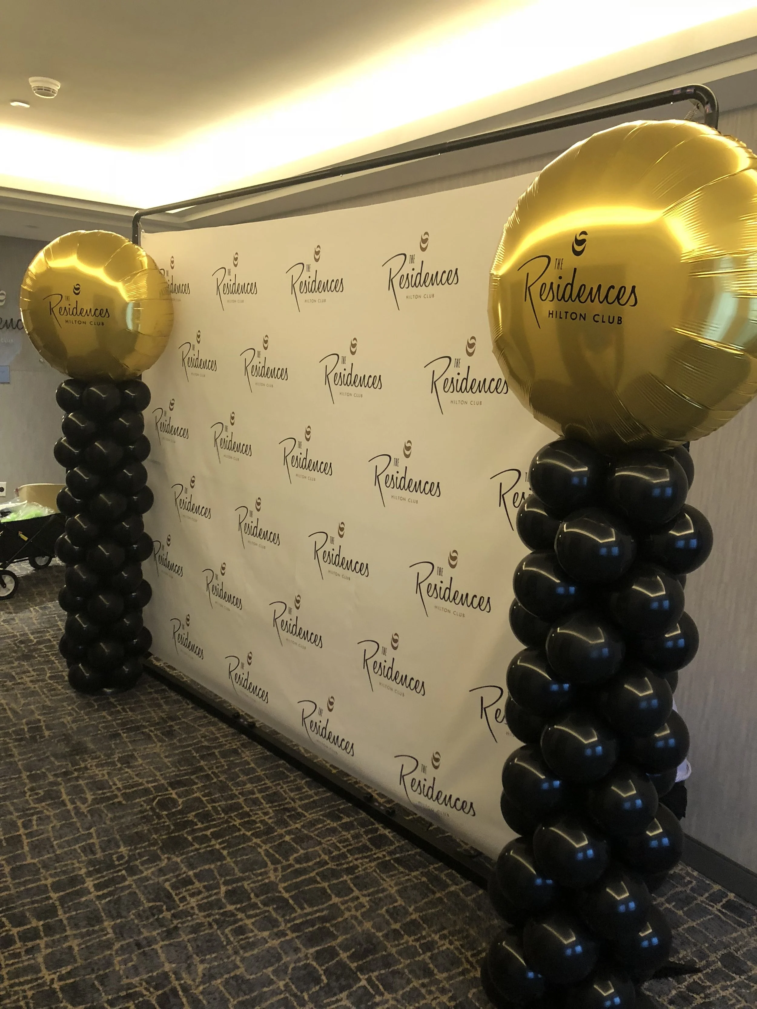 Decorative event backdrop with two balloon columns topped with gold foil balloons, featuring the logo and name of The Residences Hilton Club, set up in an indoor venue.