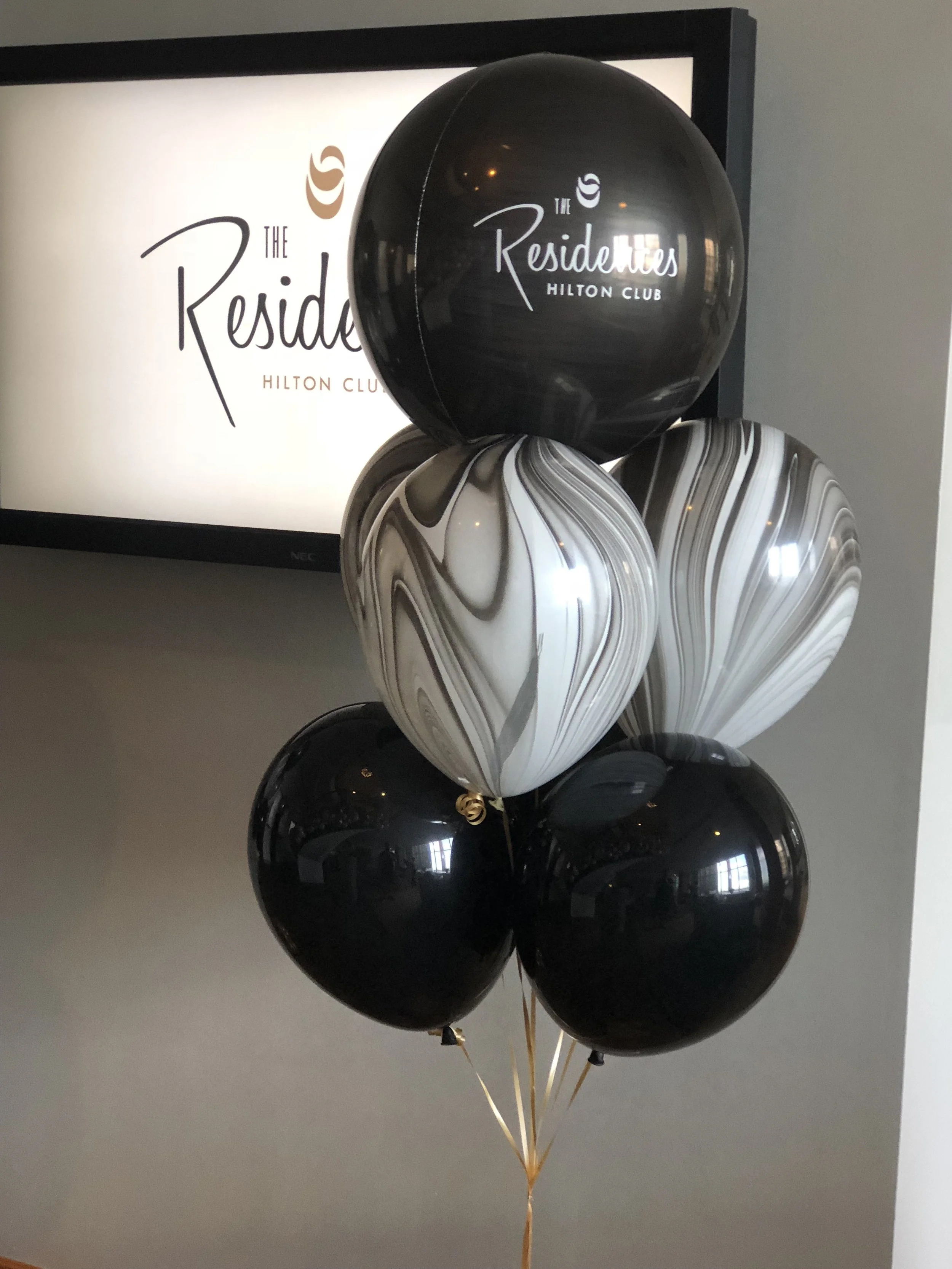 A bunch of black, white, and marbled balloons in front of a large television screen. The balloons read 'The Residences Hilton Club'.