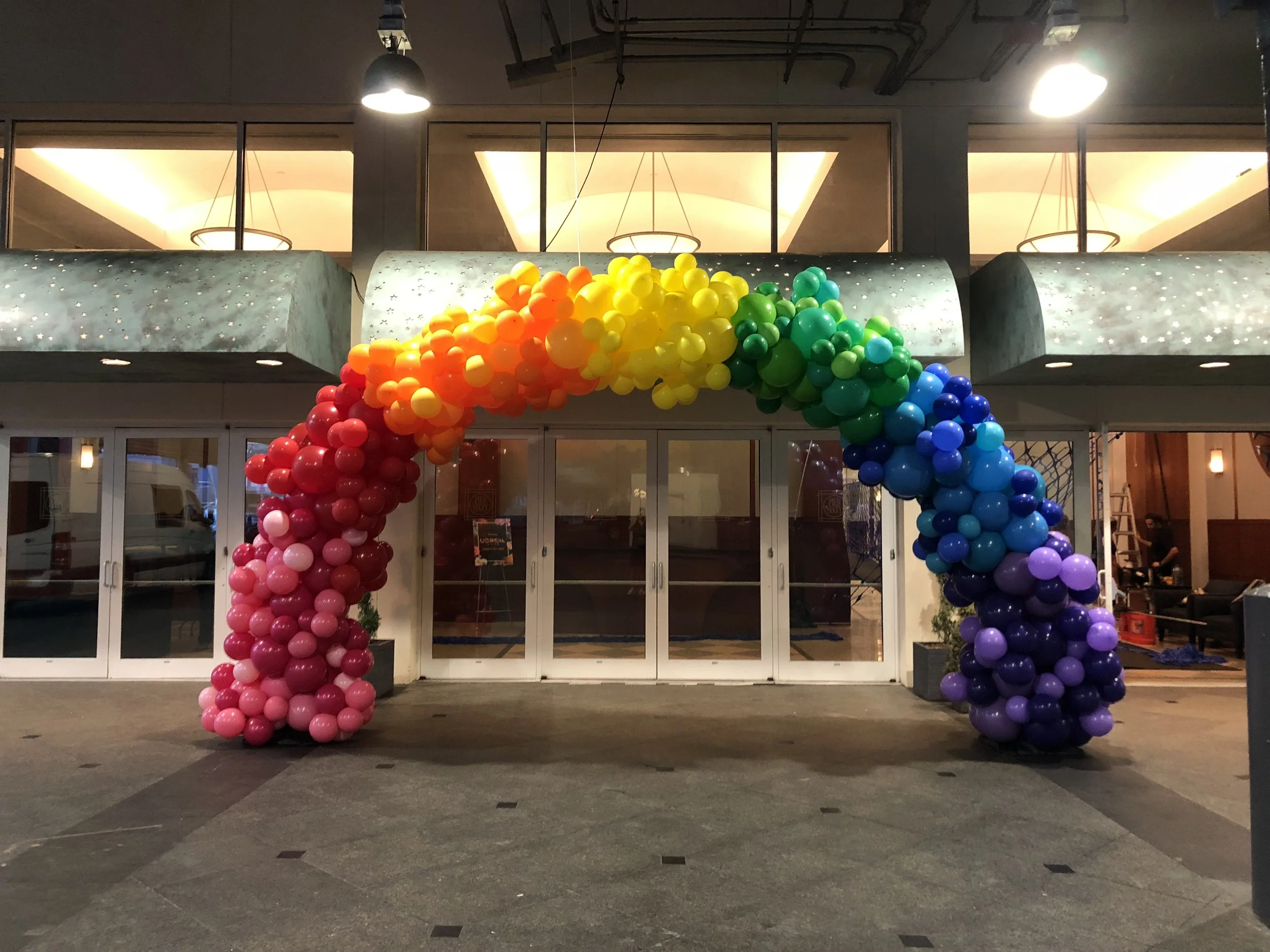 Rainbow balloon arch with red, orange, yellow, green, blue, purple clusters outside an entrance with glass doors.