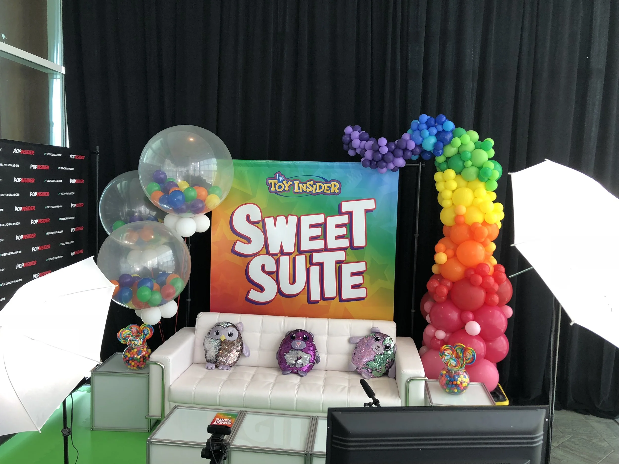 Colorful Toy Insider Sweet Suite event setup with a rainbow of balloons, plush toys, white couch, bright backdrop, and professional lighting equipment.