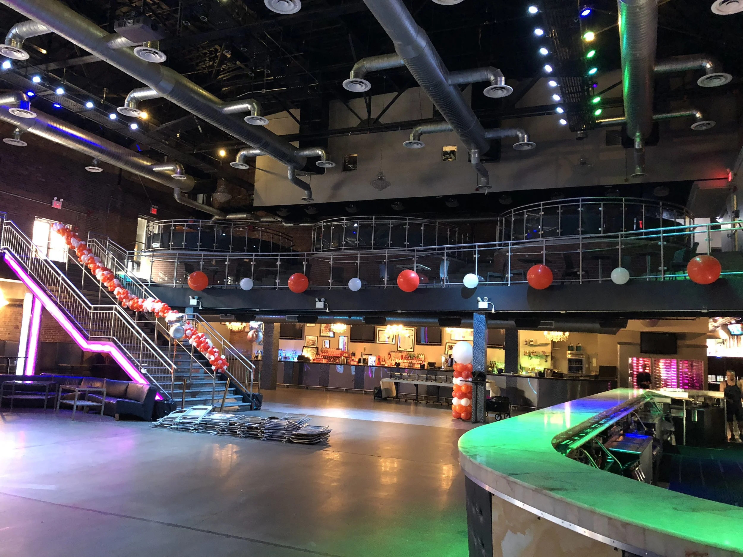 Interior of a nightclub decorated with balloons in red, white, and silver, featuring a staircase with pink neon lights, a bar area, and a spacious dance floor with colorful lighting.