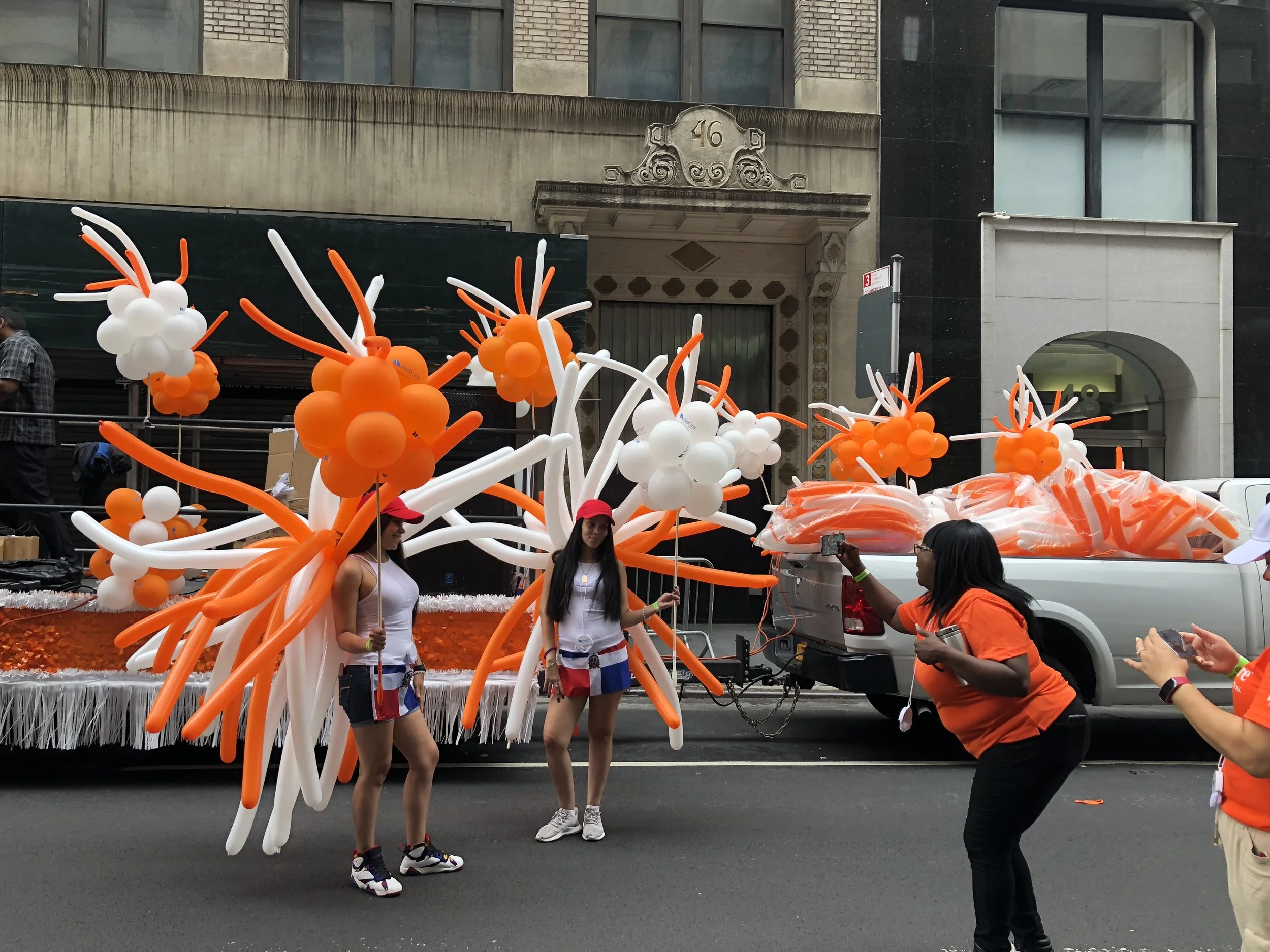 People standing in front of a float with large orange and white balloon decorations, some taking photos during a parade.