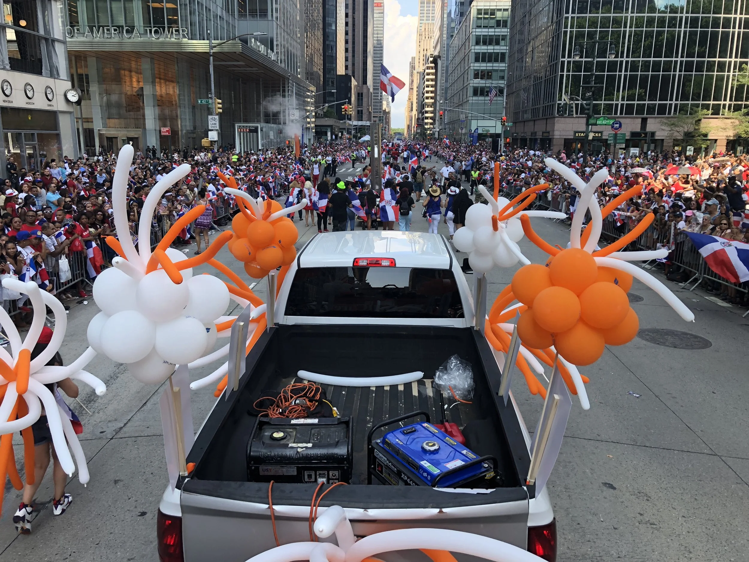 A parade with a float decorated with white and orange balloons shaped like flowers, moving down a city street crowded with people holding flags.