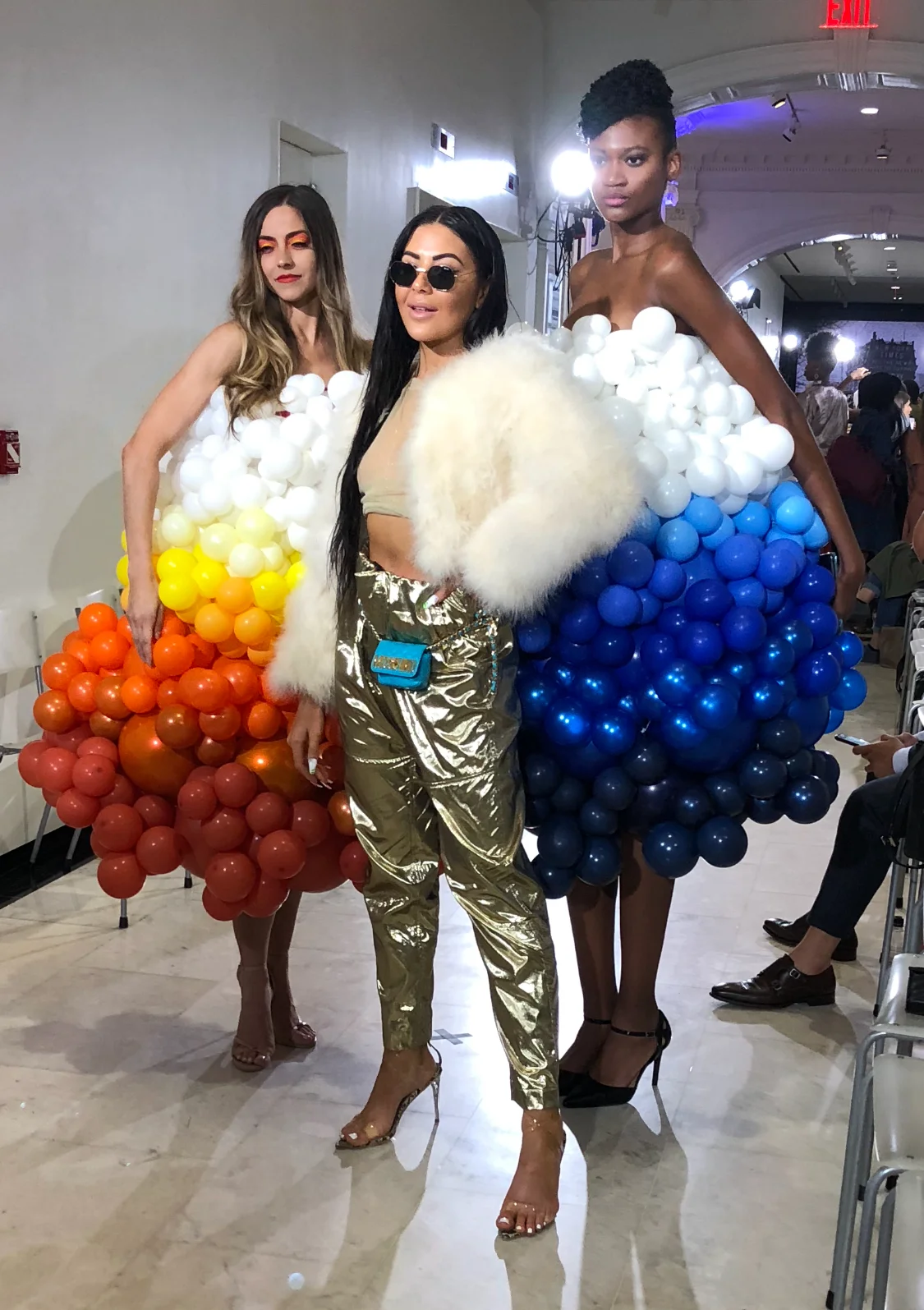 Three women posing at at a NYC Fashion week event, two models dressed in dresses made of colorful balloons and one model in metallic pants with a fur stole.