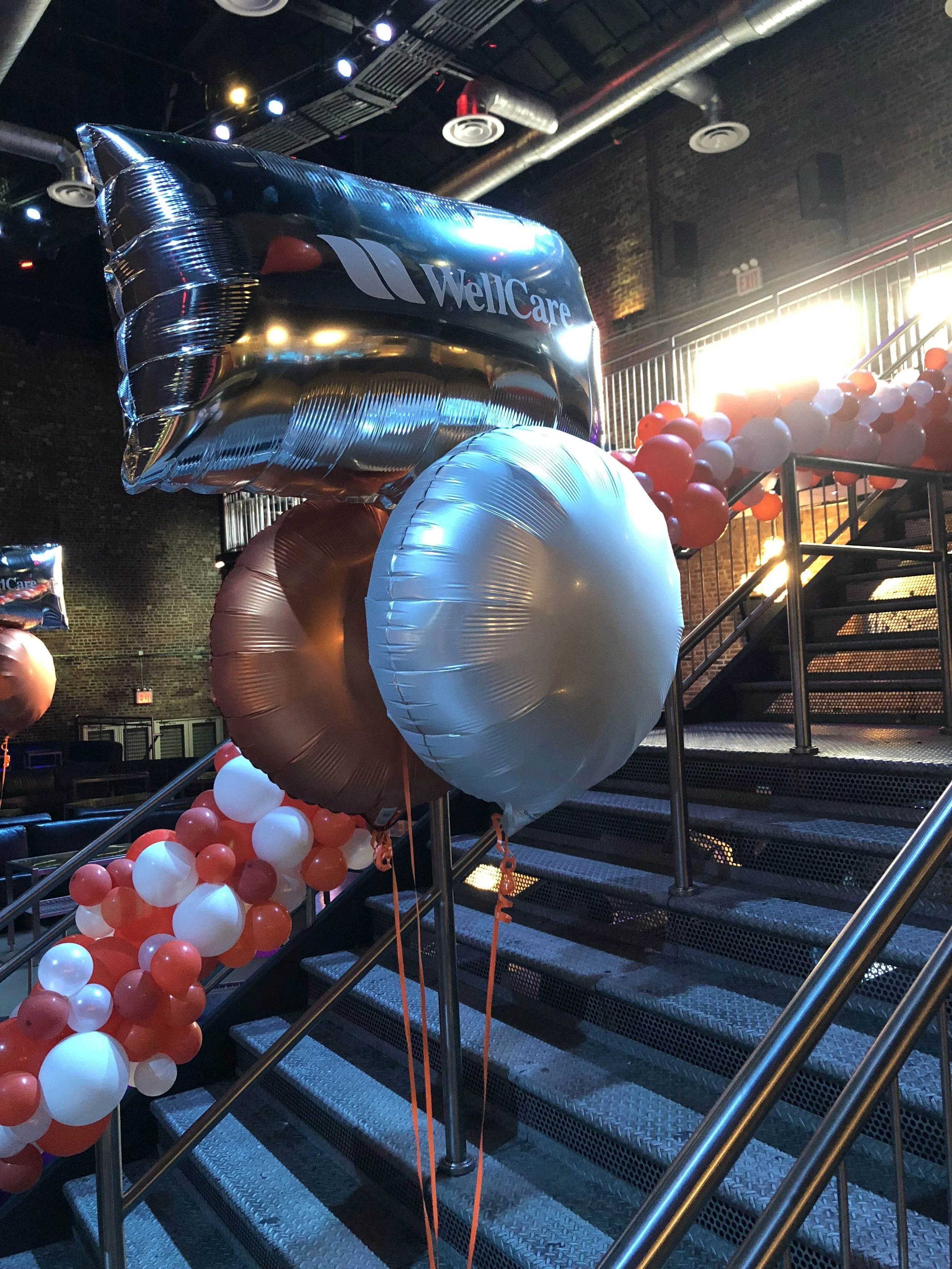 Celebration with large balloons, including a helium balloon with the WellCare logo, and red, white, and gold balloons on a staircase inside a decorated venue.