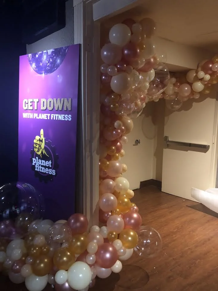 Decorative balloon arch with pink, gold, white, and transparent balloons at the entrance of a Planet Fitness gym, alongside a purple sign with a globe and a thumbs-up hand, advertising 'Get Down with Planet Fitness'.