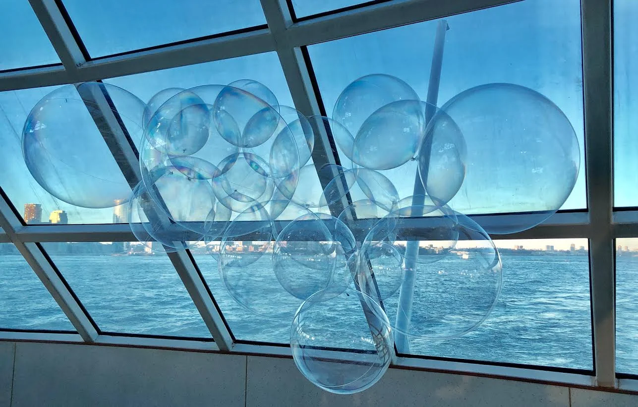 Art installation of transparent balloons inside a glass-paneled room overlooking a body of water and a city skyline in the distance.