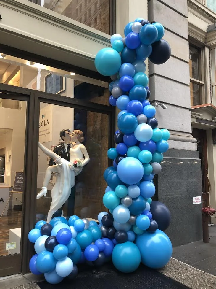 A storefront decorated with blue, teal, black, and grey balloons in various sizes surrounding a window display of a wedding-themed statue of a bride and groom kissing.
