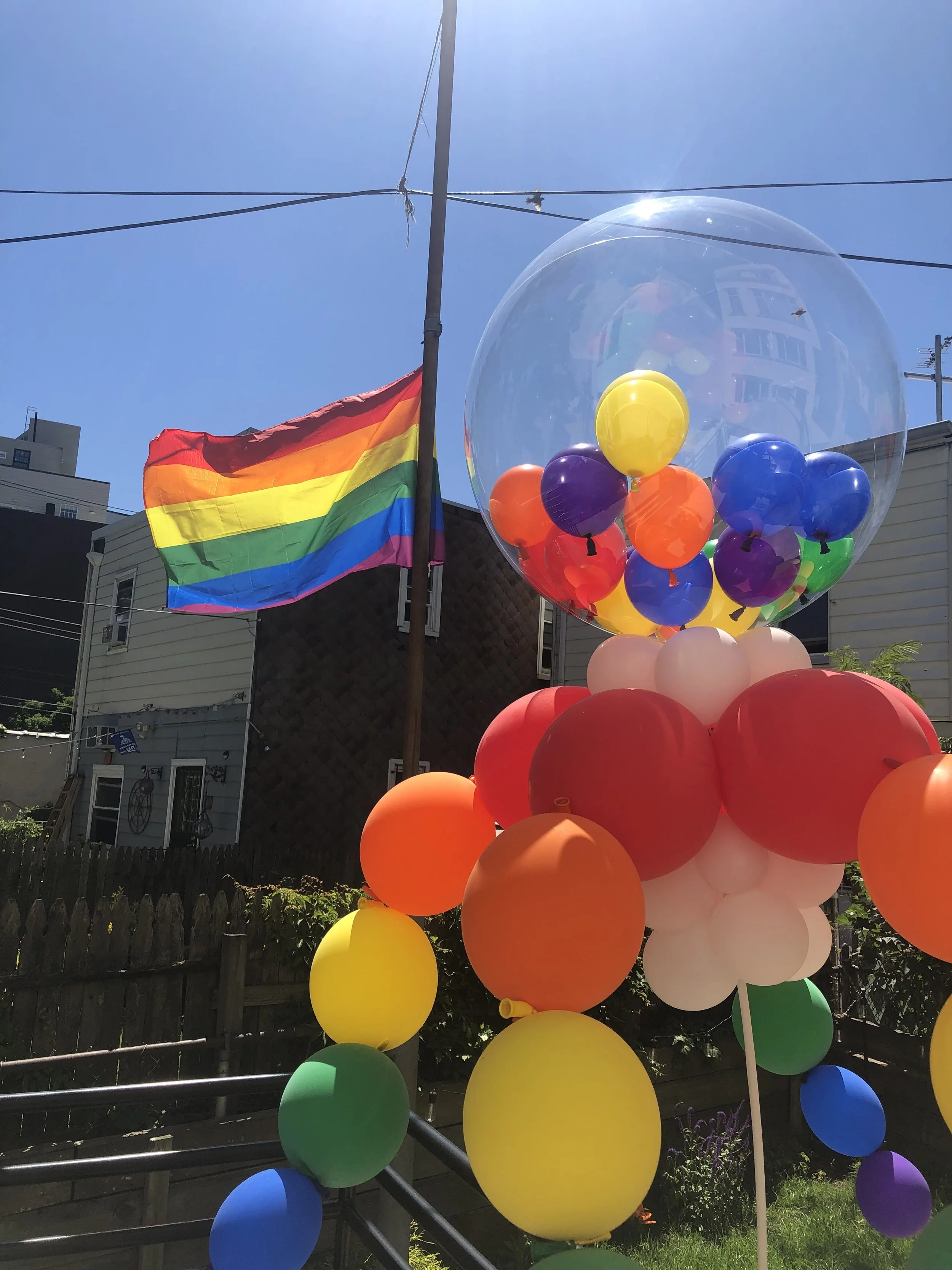 Colorful balloons arranged in a sculpture with a rainbow flag flying in the background under a clear blue sky.